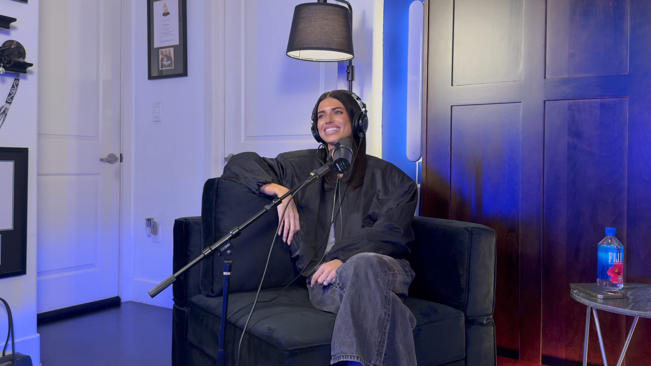A young woman is sitting on a black couch wearing headphones and speaking into a microphone during a podcast recording. She is smiling and dressed in a black jacket and gray pants. Behind her, there is a wooden wall, a standing lamp, and framed pictures on the white wall. A water bottle is placed on a small round table next to her.