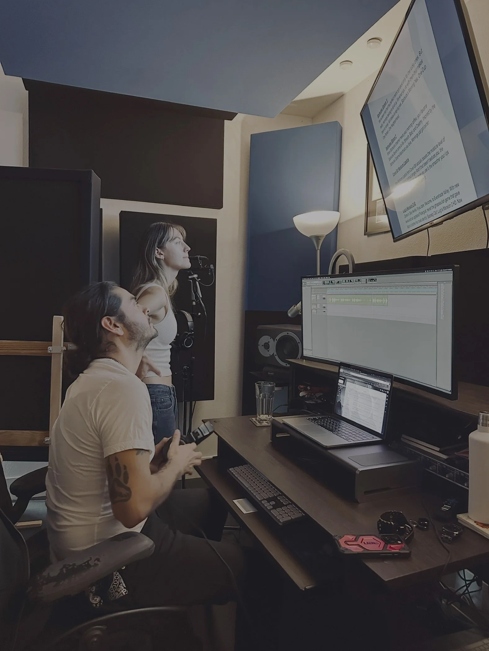 People in a recording studio working with computers, audio equipment, and monitors, with acoustic panels on the walls.