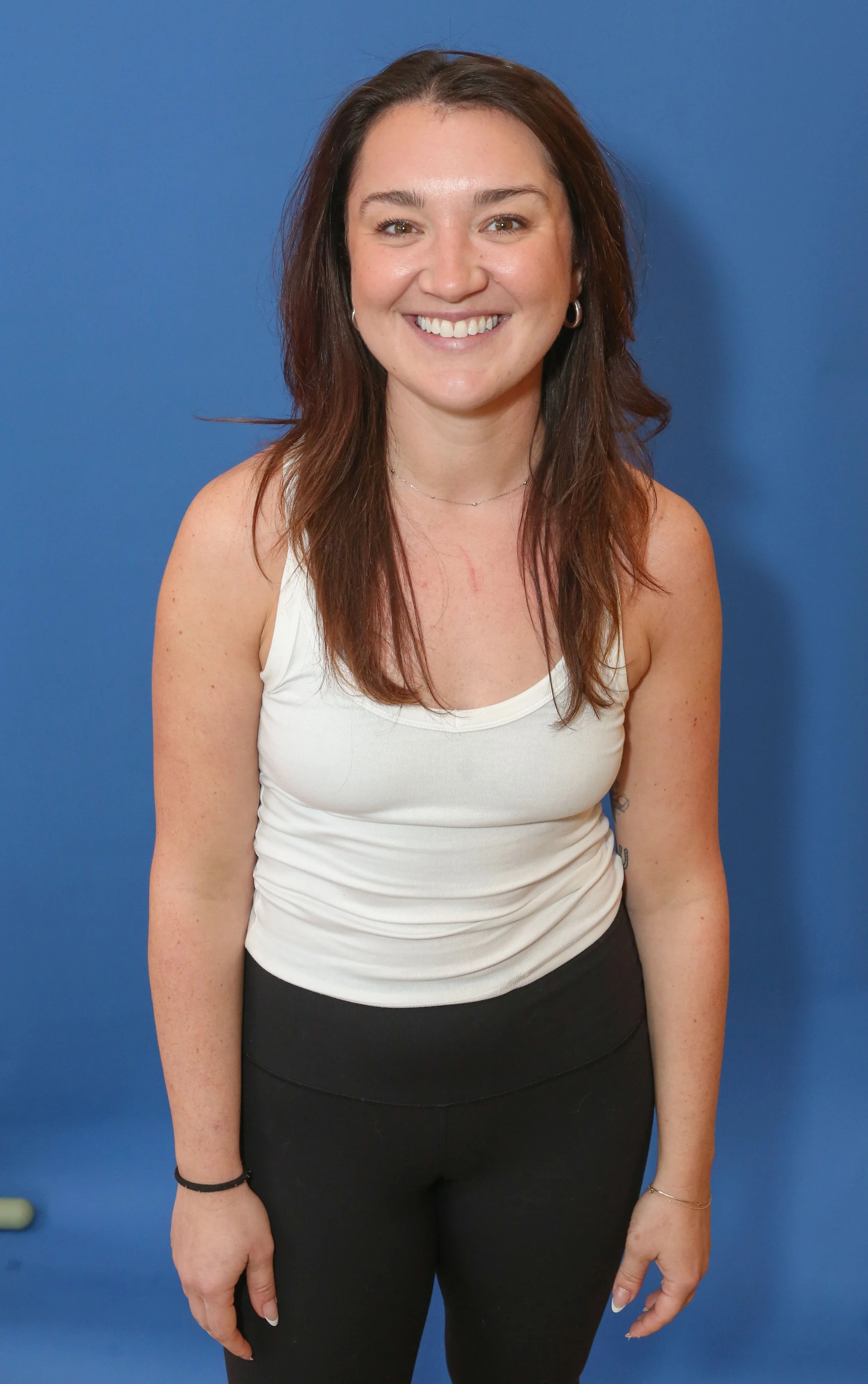 A woman with brown hair holding two light blue dumbbells, smiling, standing against a pink background.