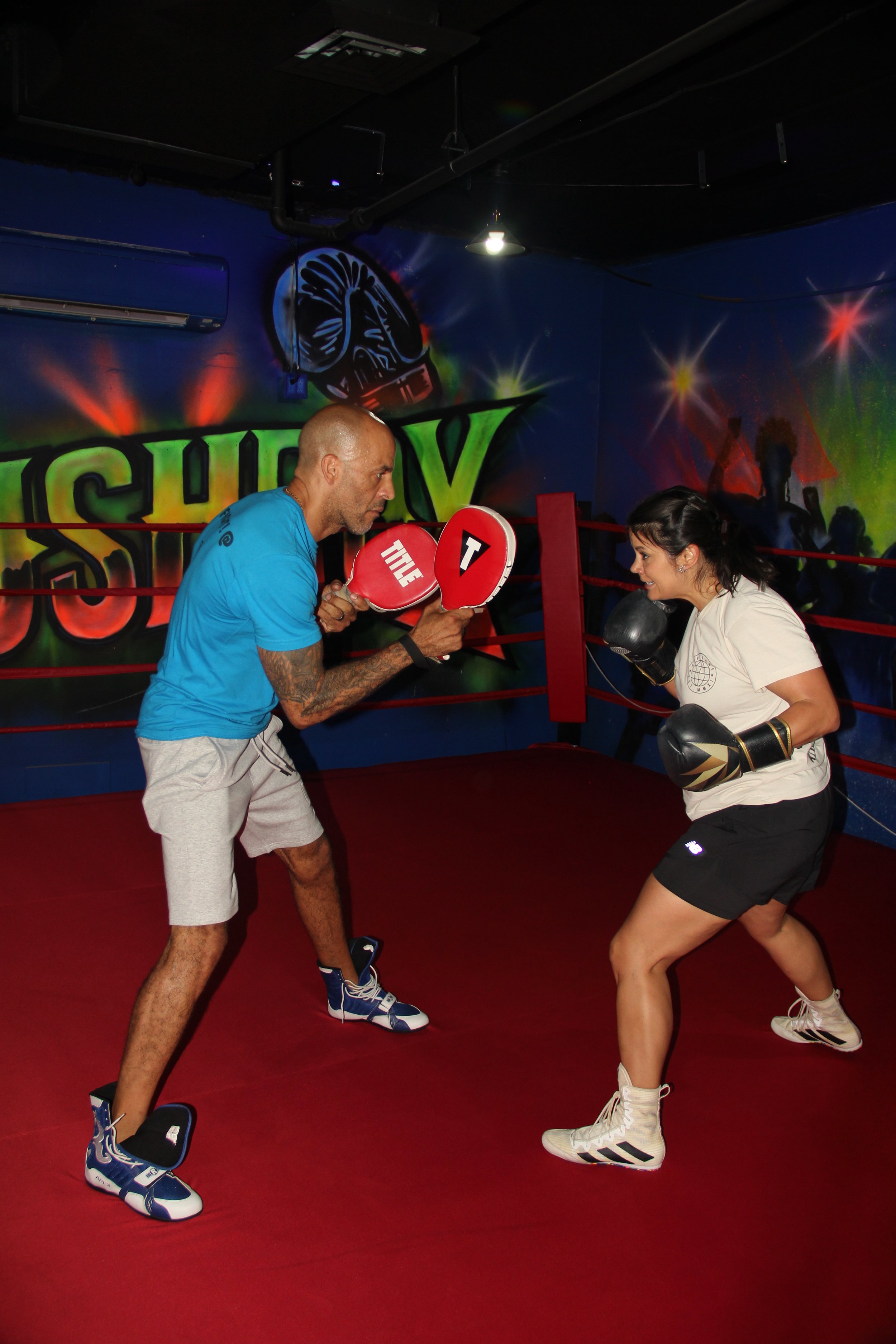 A man and a woman sparring in a boxing gym, with the man holding focus mitts and the woman wearing boxing gloves, in a boxing ring with red ropes and a colorful graffiti wall in the background.