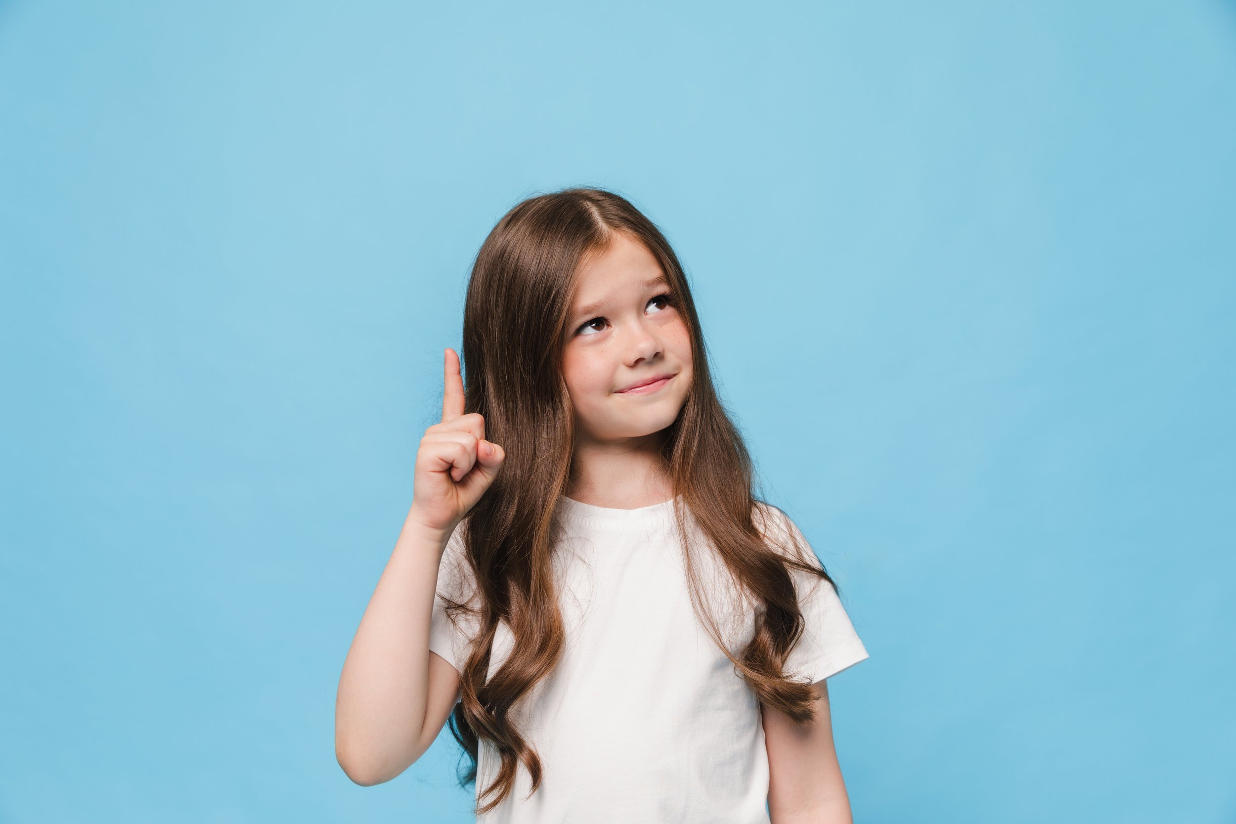 A young girl with long brown hair pointing her finger upward with a thoughtful expression against a light blue background, representing a child's curiosity in inquiry-based learning.