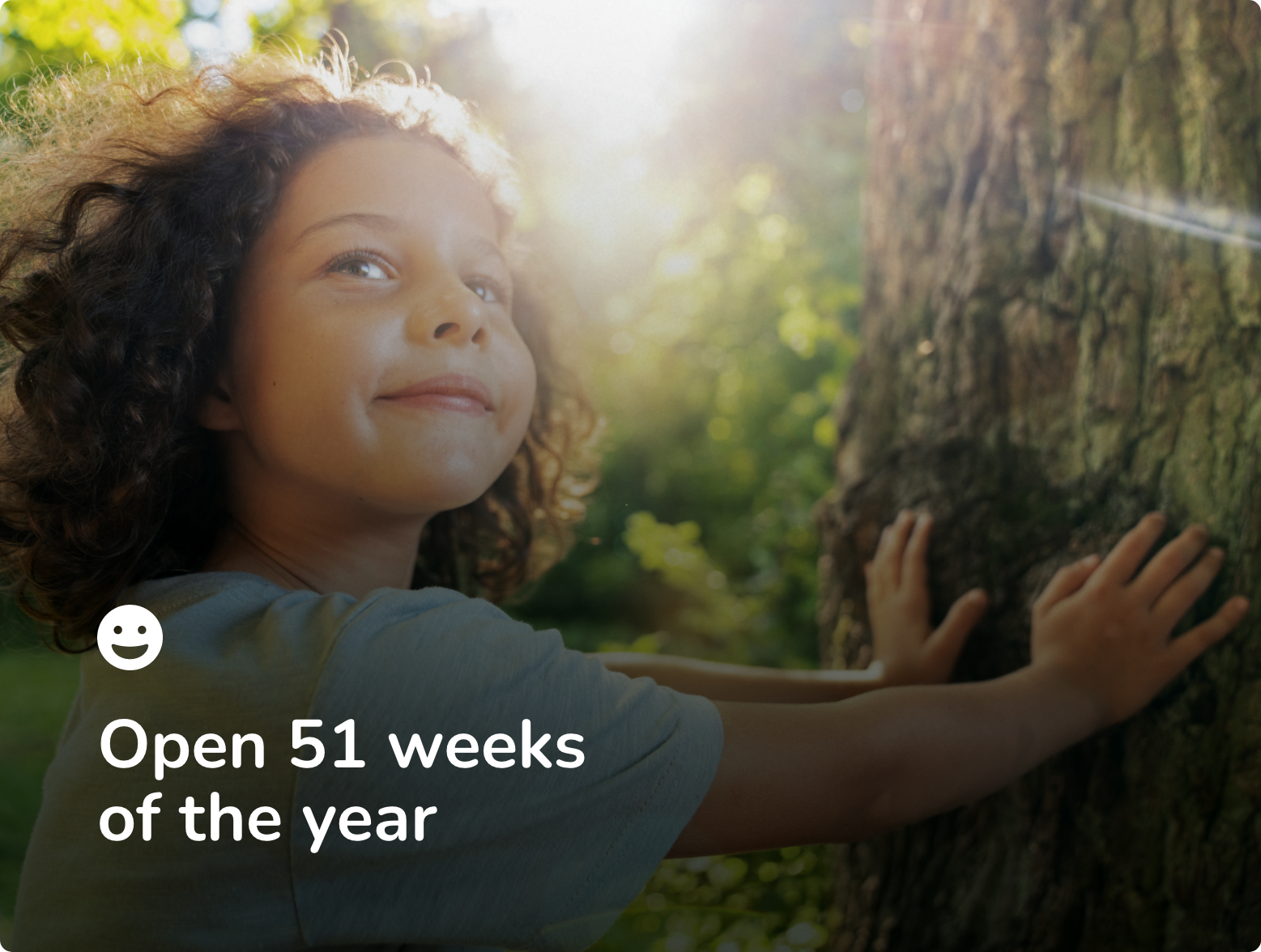 A young girl with curly hair hugging a tree in a forest, sunlight shining behind her, with text overlay that reads 'Open 50 weeks of the year'.