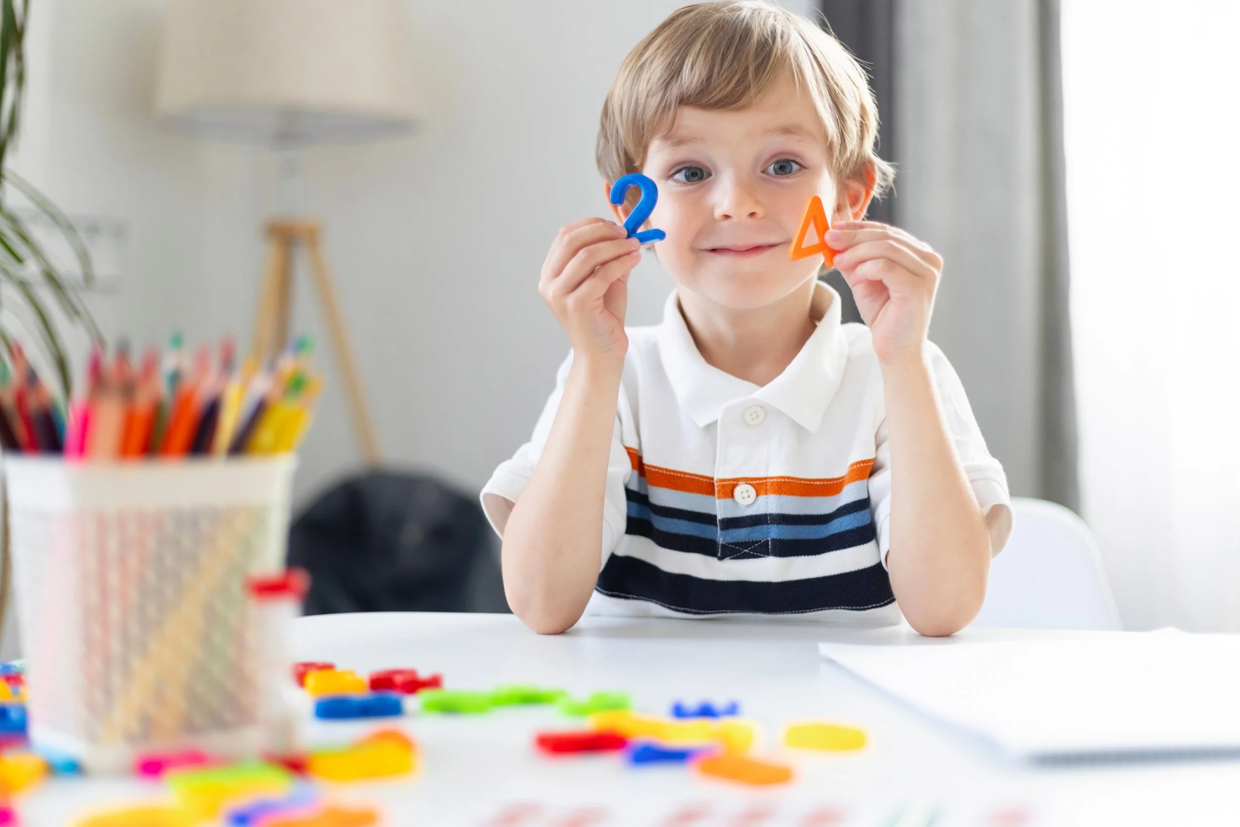 A smiling young boy holding up colourful plastic numbers at a desk, engaging in hands-on early learning.