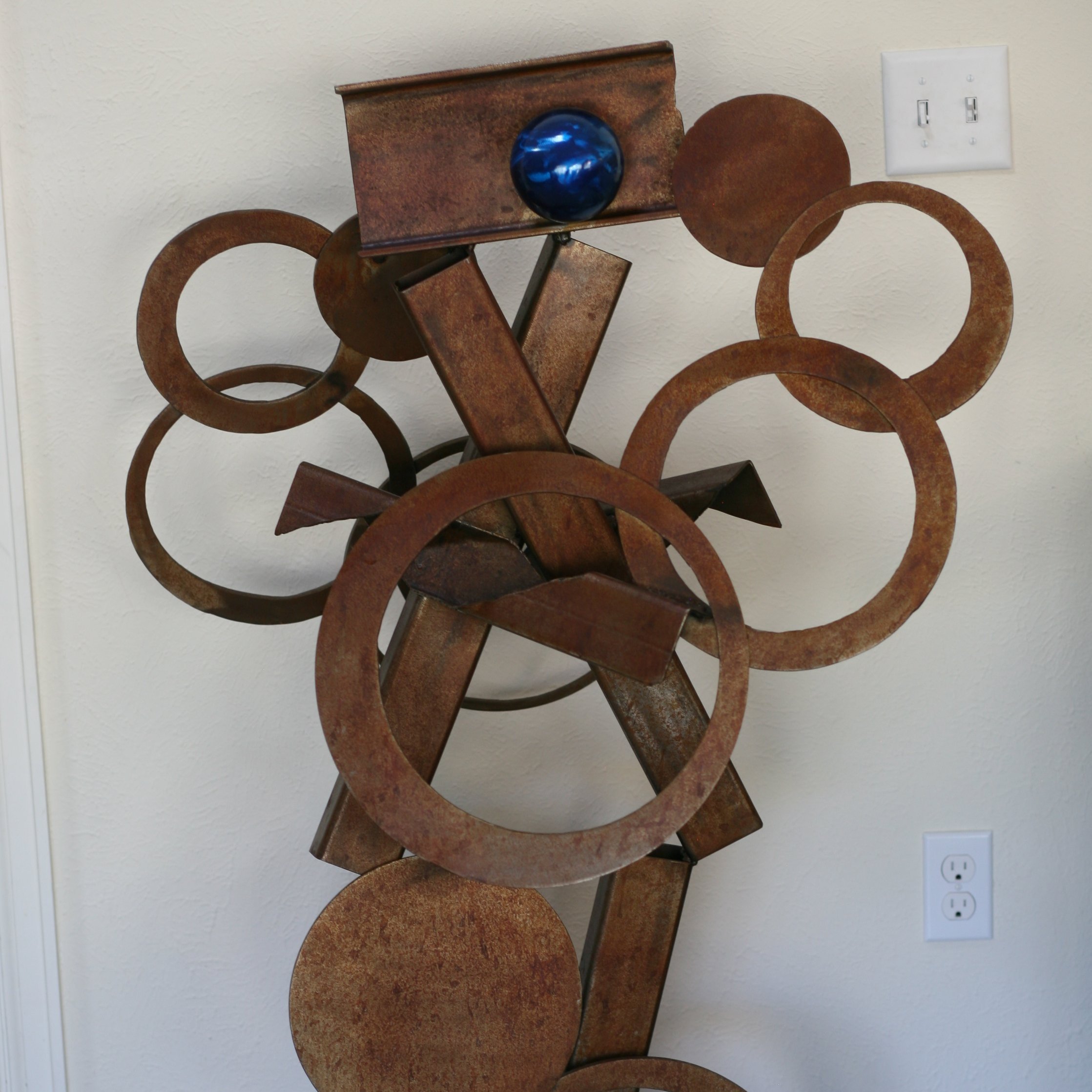 Rusted steel abstract sculpture with I-beam, rings, and tubing against a wall