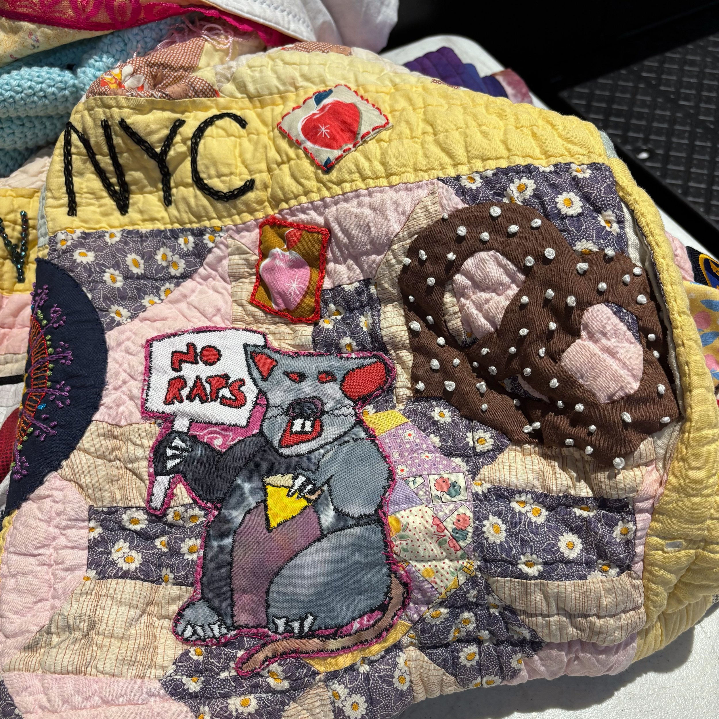 Close-up of a quilt with various pasted patches including the words 'MY CAT RAPS' embroidered in black on a yellow patch, a red heart with a tag, and other colorful fabric designs with floral patterns, pearls, and embroidered elements.