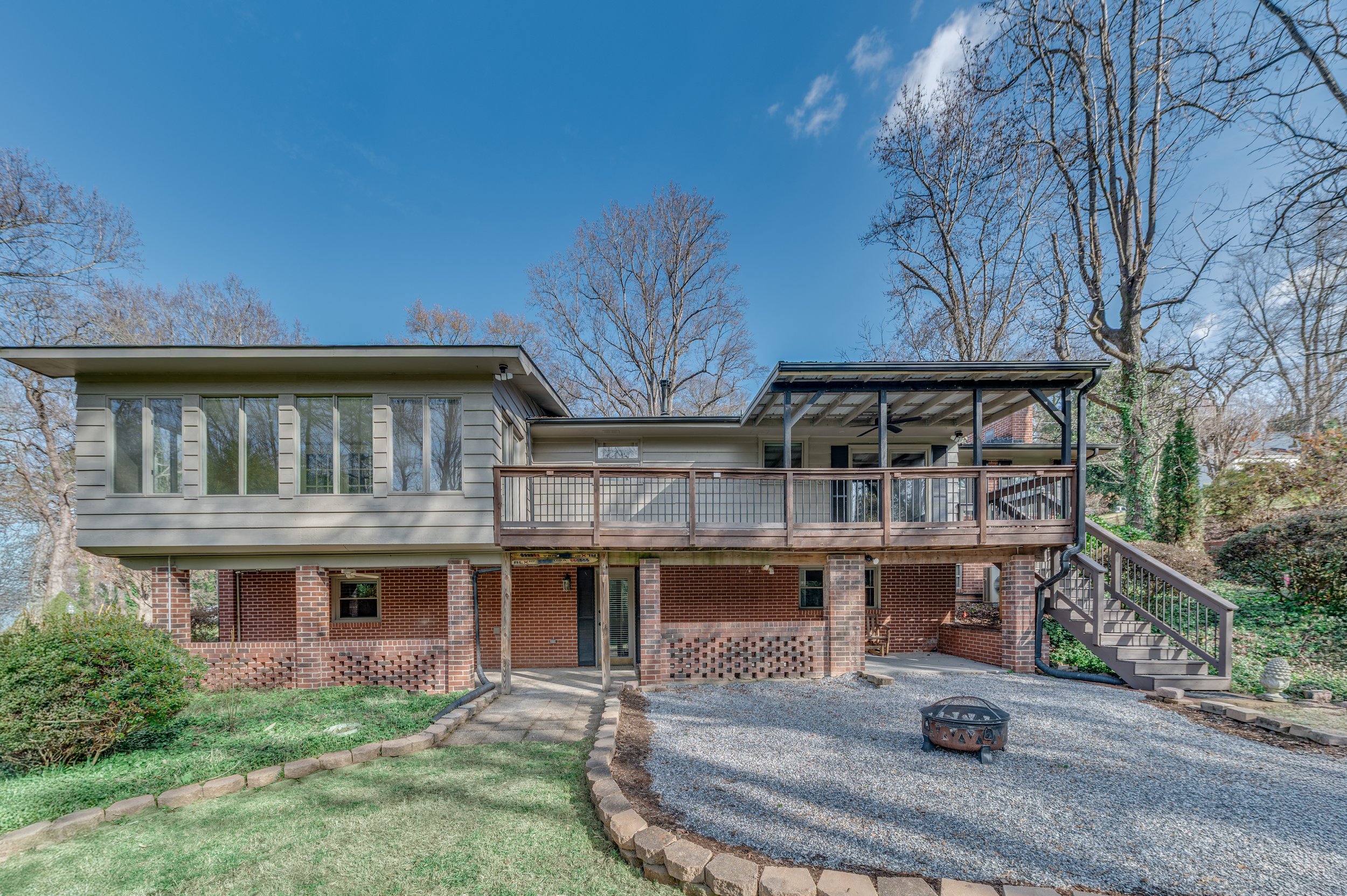A two-story house with a brick basement and a wooden upper floor. The house has large windows, a raised deck with a railing, a staircase leading up to the deck, and a gravel driveway with a fire pit in the yard. The background features leafless trees