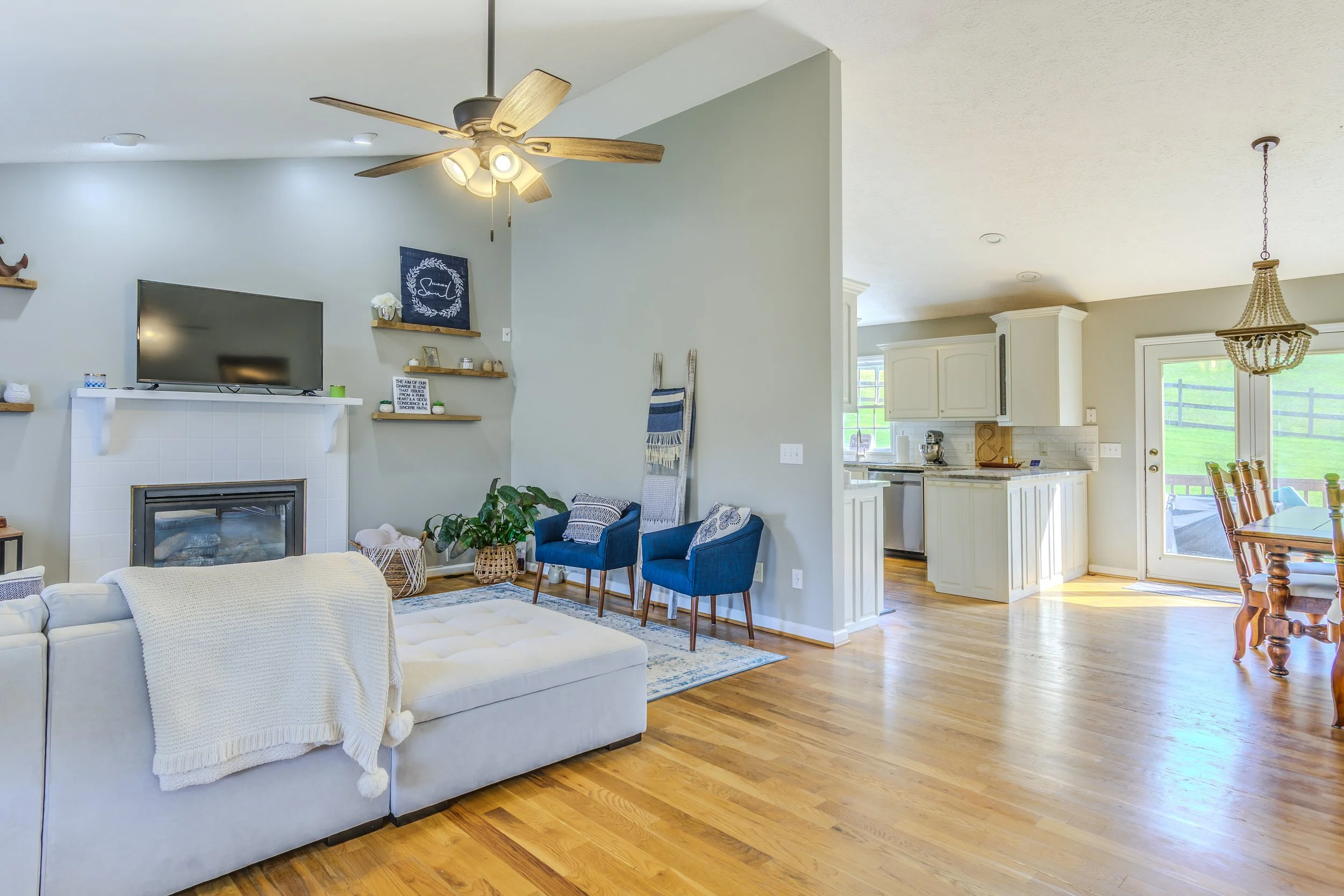 Open-concept living room and kitchen with hardwood floors, a ceiling fan, a fireplace, a wall-mounted TV, blue accent chairs, a dining area, and sliding glass door leading outside.