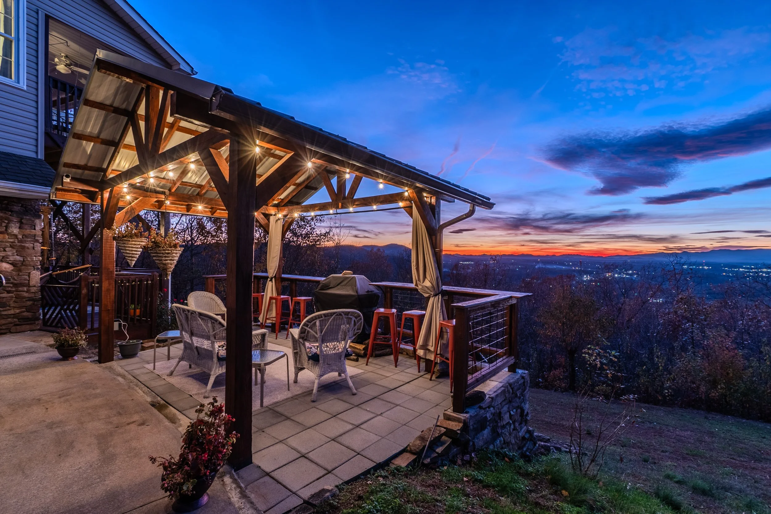 Outdoor patio with string lights, chairs, and a barbecue grill, overlooking a sunset and distant mountains.