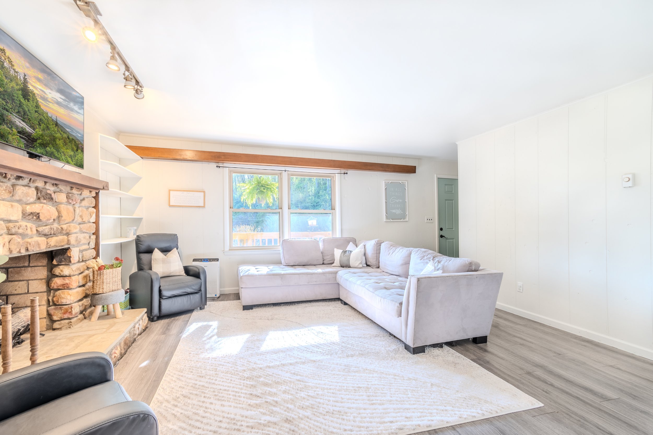 Bright living room with large beige sectional sofa, black armchair, fireplace, and window allowing natural light, with light-colored walls and wooden accents.