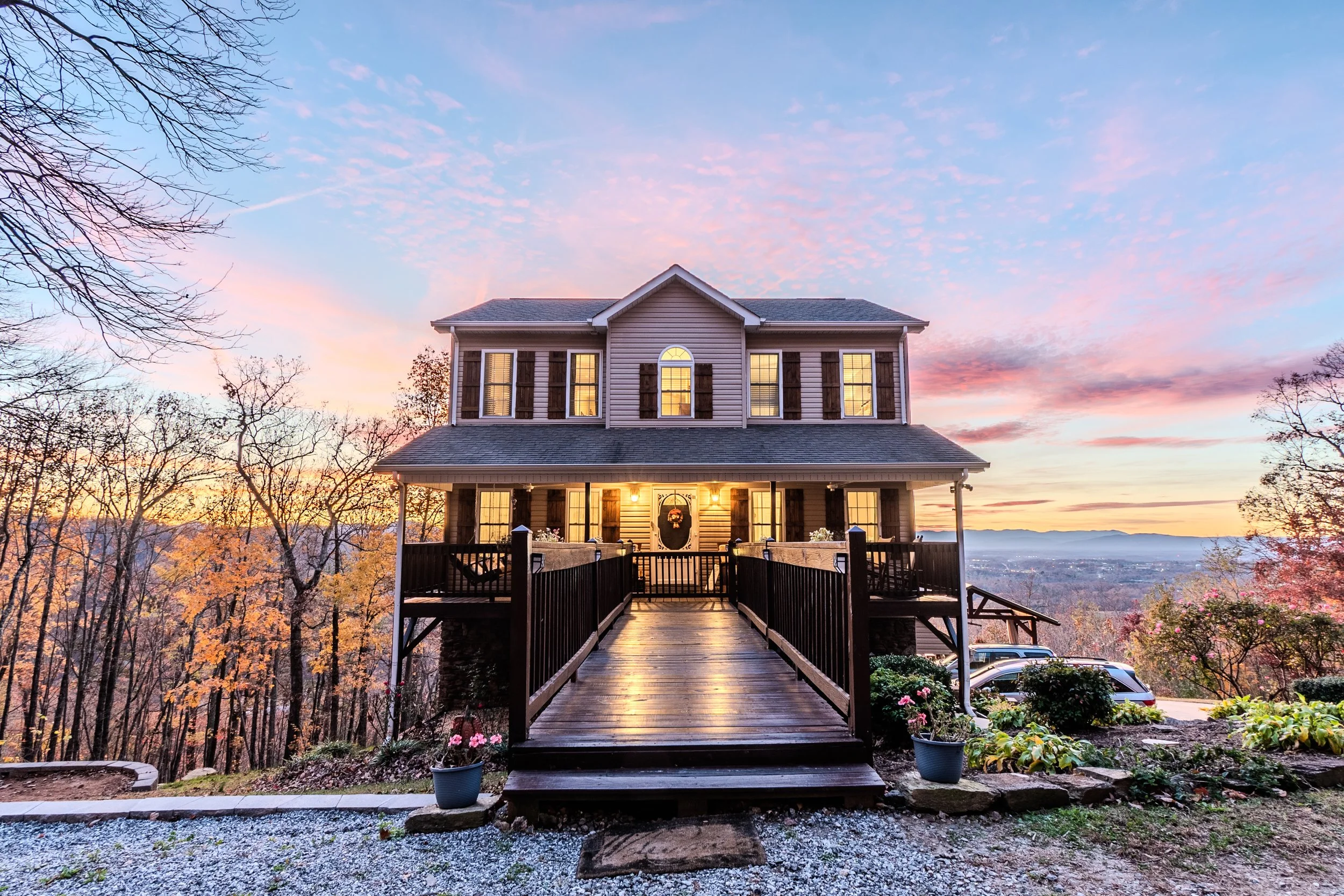 A two-story house with a wrap-around porch, wooden stairs, and warm outdoor lighting, set against a colorful sunset sky with trees and distant mountains.