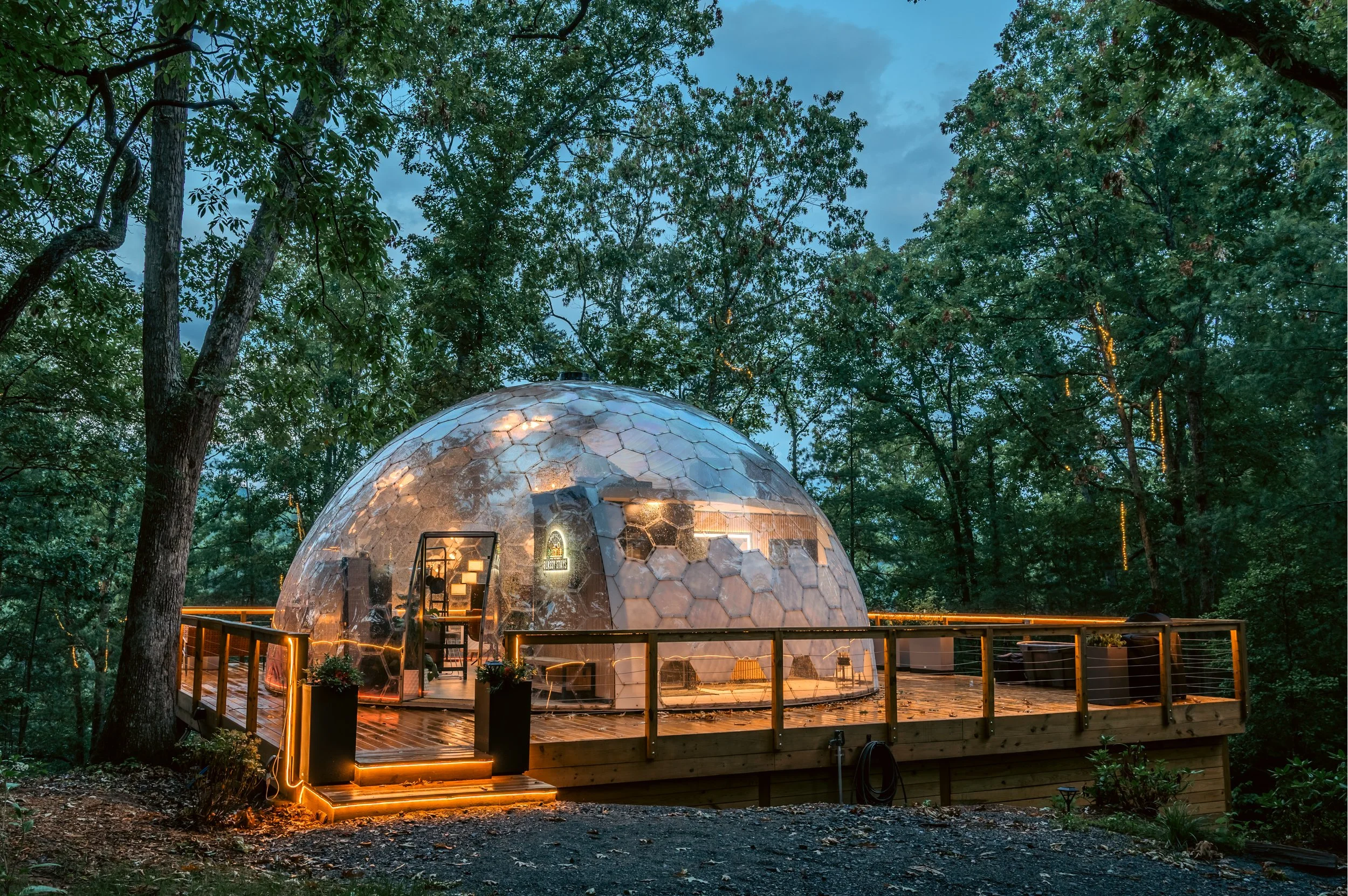 A geodesic dome house with transparent walls and wood deck, surrounded by trees at dusk, with warm lighting inside and on the deck.