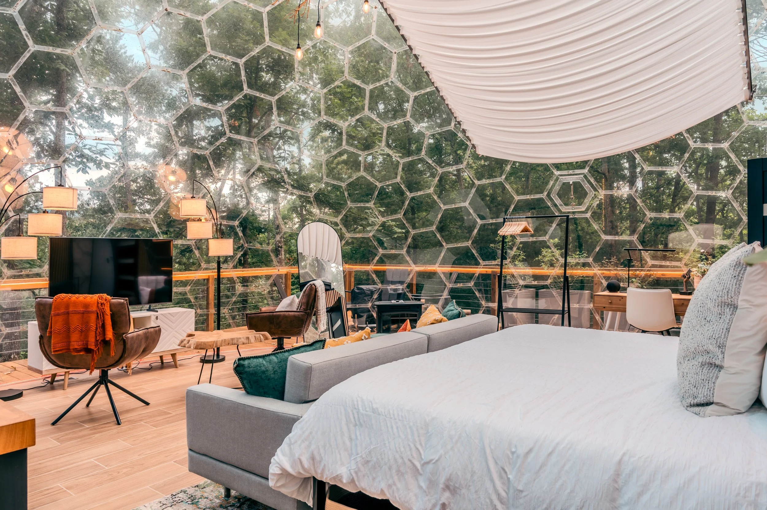 A modern bedroom with a large bed covered in white bedding, a gray sofa, and a geometric glass wall looking out to trees. The room features a wooden floor, a flat-screen TV, a brown chair with an orange throw blanket, a standing mirror, and various l