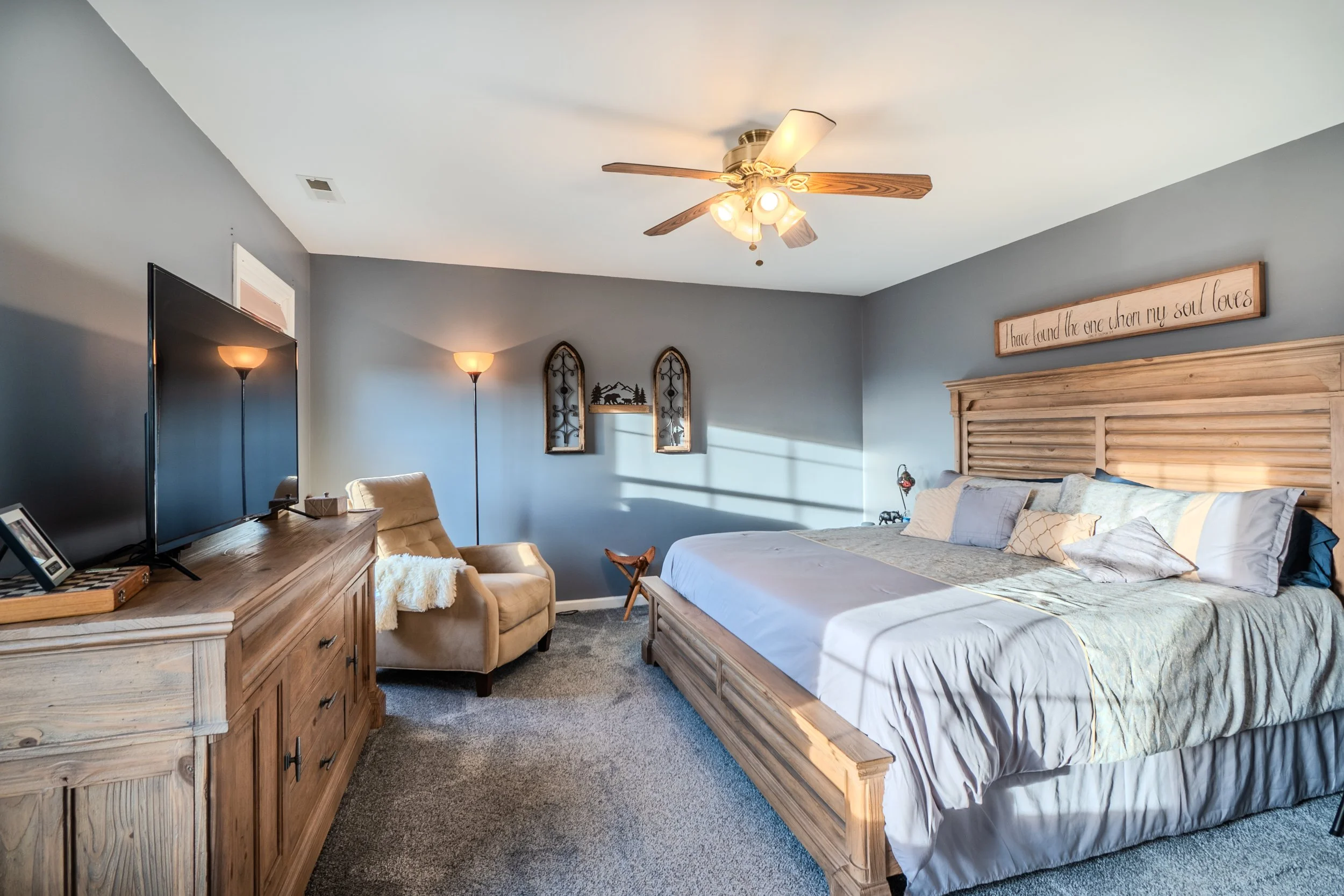 A bedroom with a large wooden bed, gray walls, a ceiling fan, a flat-screen TV on wooden furniture, a beige armchair with a fluffy throw, two floor lamps, and decorative wall hangings.