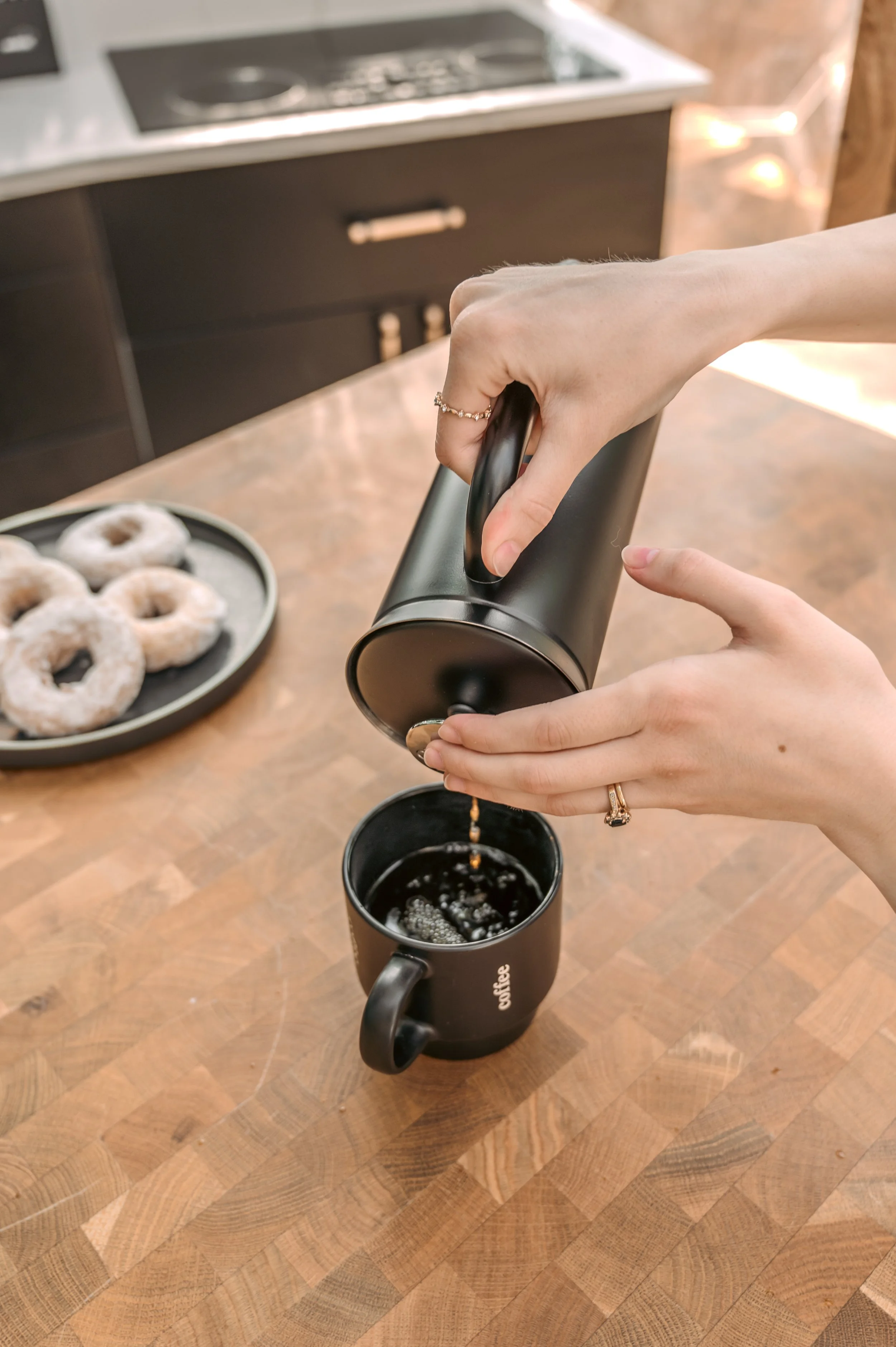 Person pouring coffee from a black French press into a black mug, with a plate of donuts in the background.