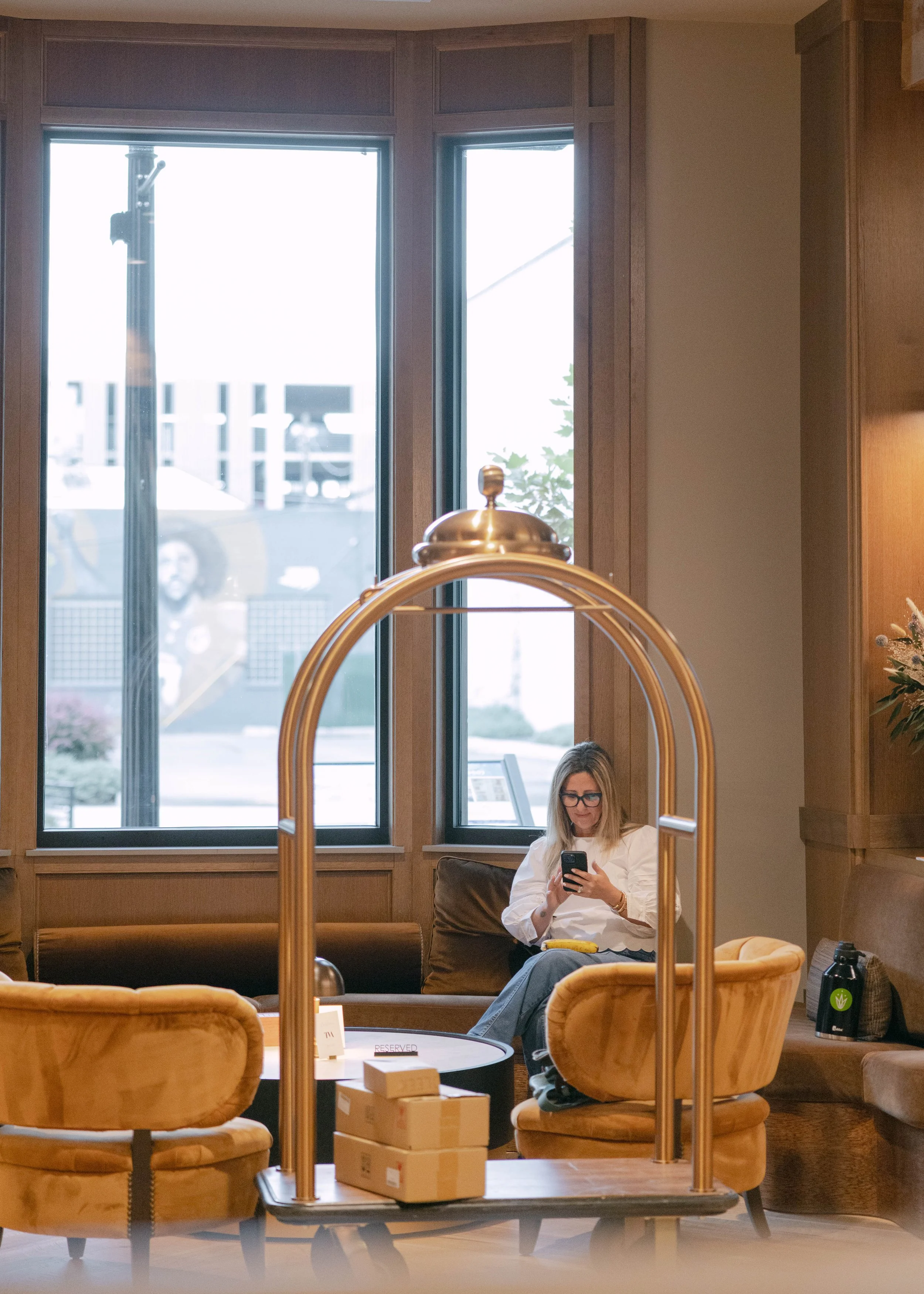 A woman with blond hair, wearing glasses, a white blouse, and jeans, is sitting on a brown couch in a hotel lobby, looking at her phone. The lobby features large windows, wooden wall paneling, a marble table with small boxes, and a brass luggage cart in front of her.