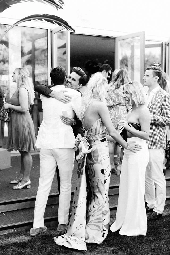 Group of people socializing at an outdoor party, some hugging and chatting on a patio with open glass doors in the background.