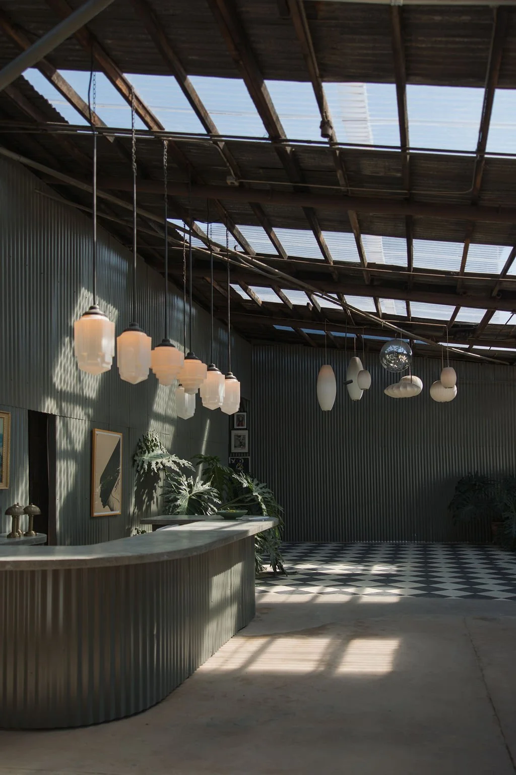 Interior of a modern building with a corrugated metal counter, hanging pendant lights, art on the wall, and large plants, with sunlight casting shadows through a partially open roof.