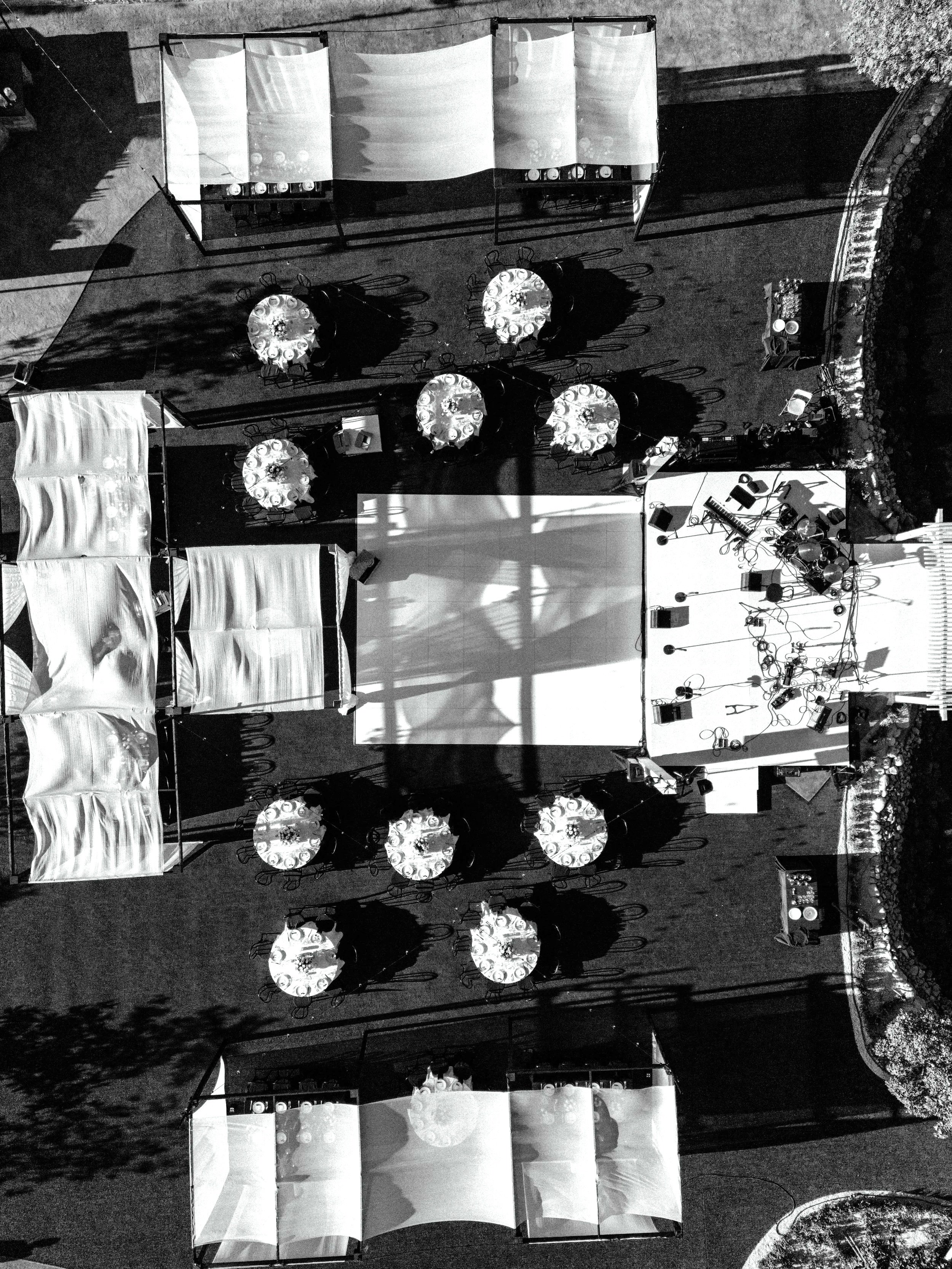 An outdoor event setup with tables and chairs, some covered with white cloth, and stage equipment including microphones, speakers, and musical instruments. Several round tables with chairs are arranged around the area, and there are shadows cast by trees or structures.