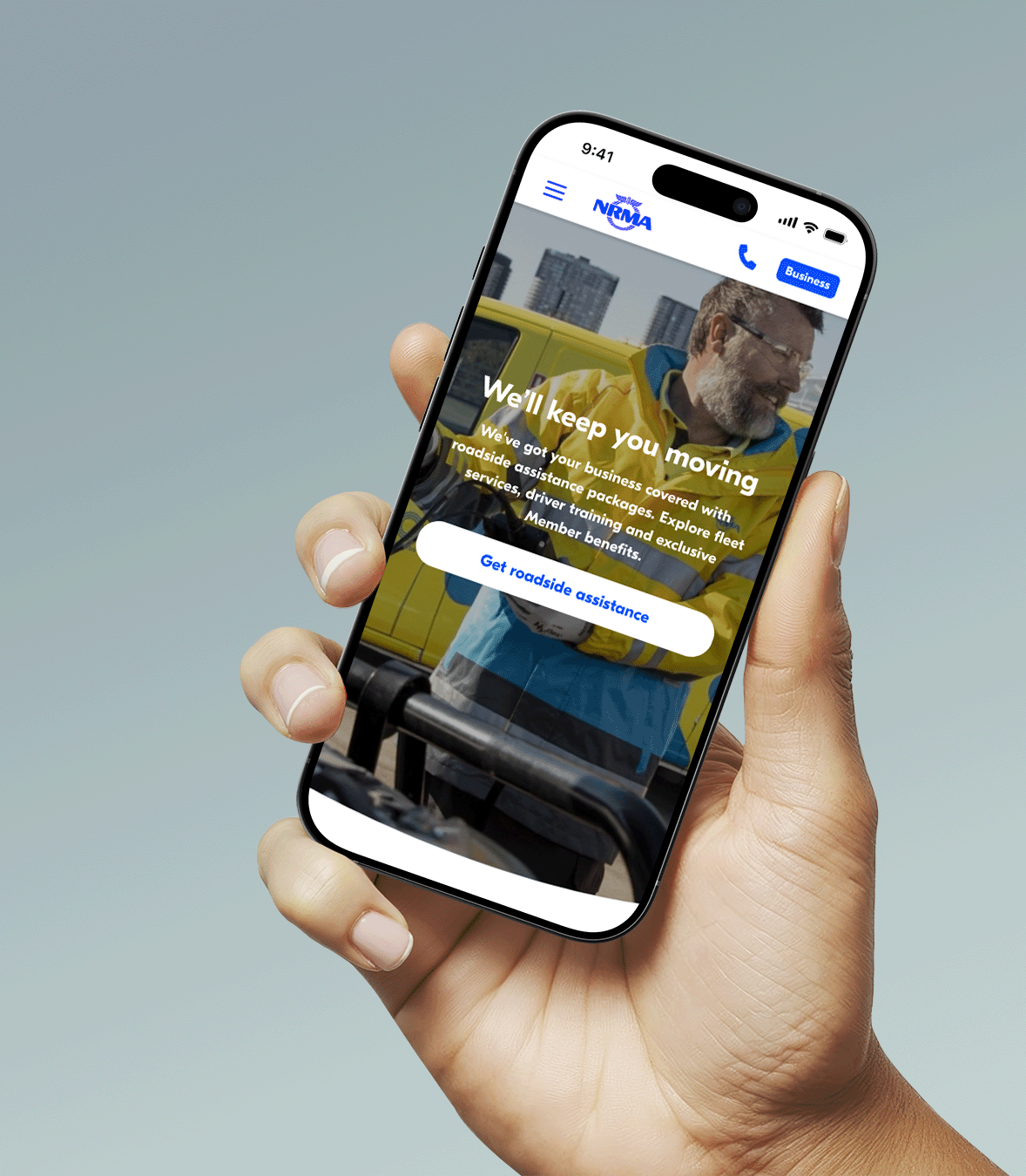 Smartphone held in a person's hand displaying a roadside assistance service app with a smiling man in a yellow jacket in front of a yellow vehicle, city buildings in the background, and a blue button inviting users to get roadside assistance.