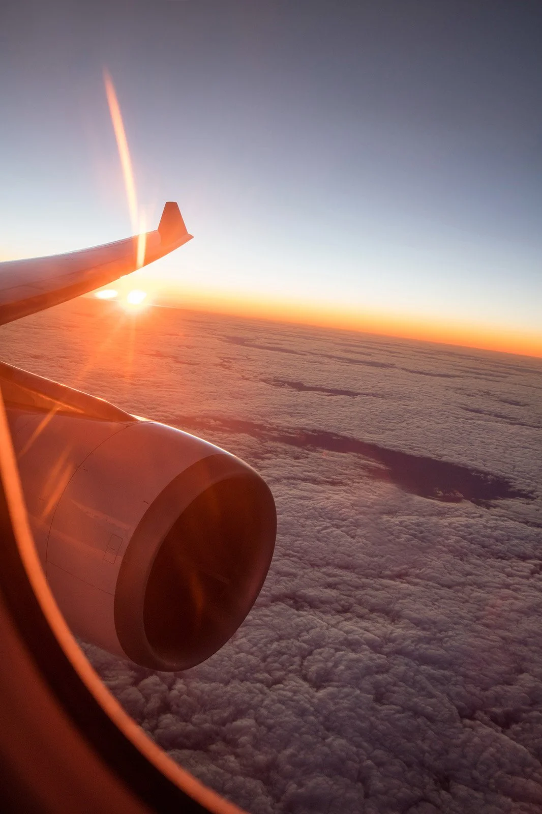 View of an airplane wing and engine during sunset, seen through the window above a layer of clouds.