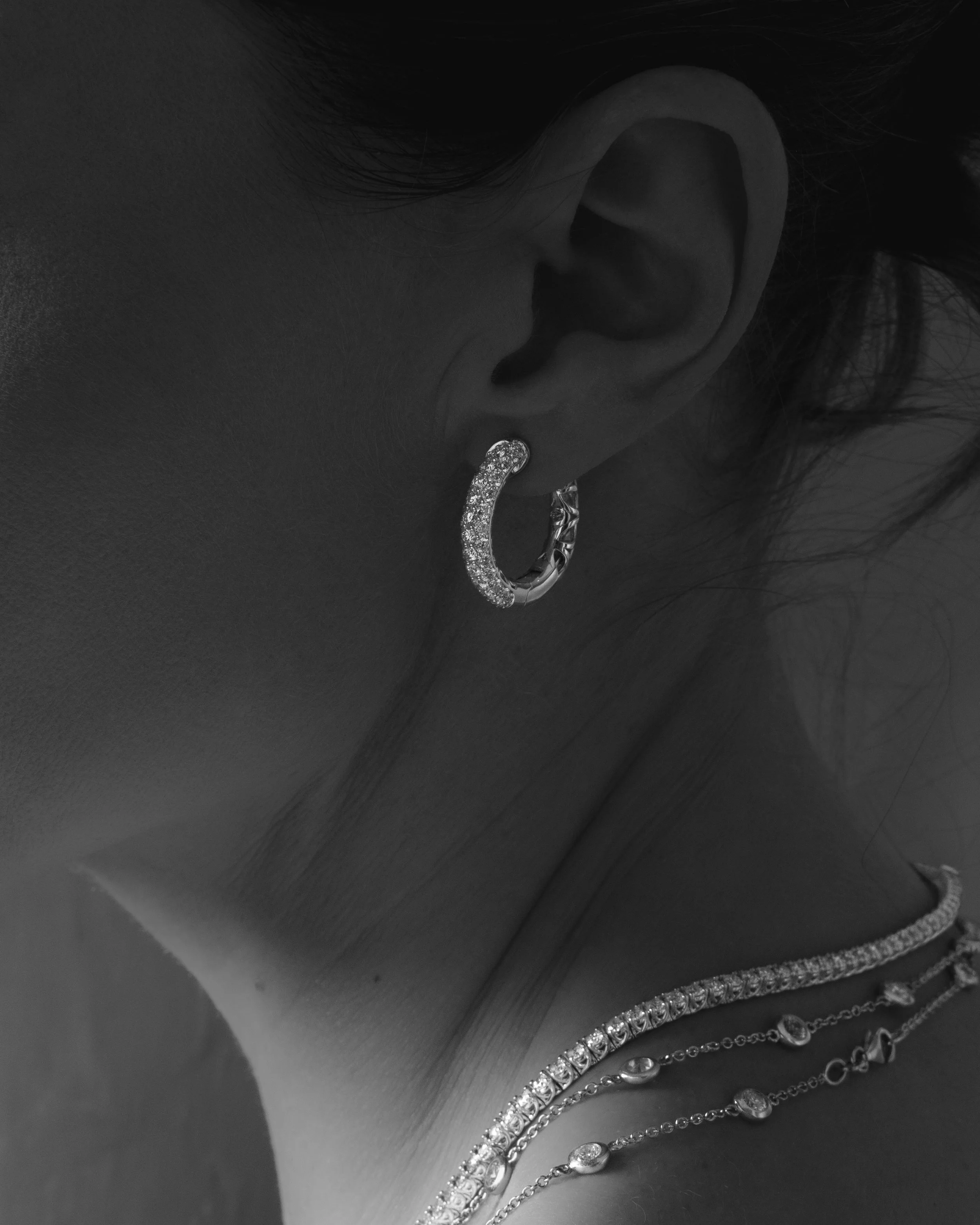 Close-up of a woman's ear and neck, showing multiple jewelry pieces including a sparkling hoop earring and layered necklaces, in black and white.