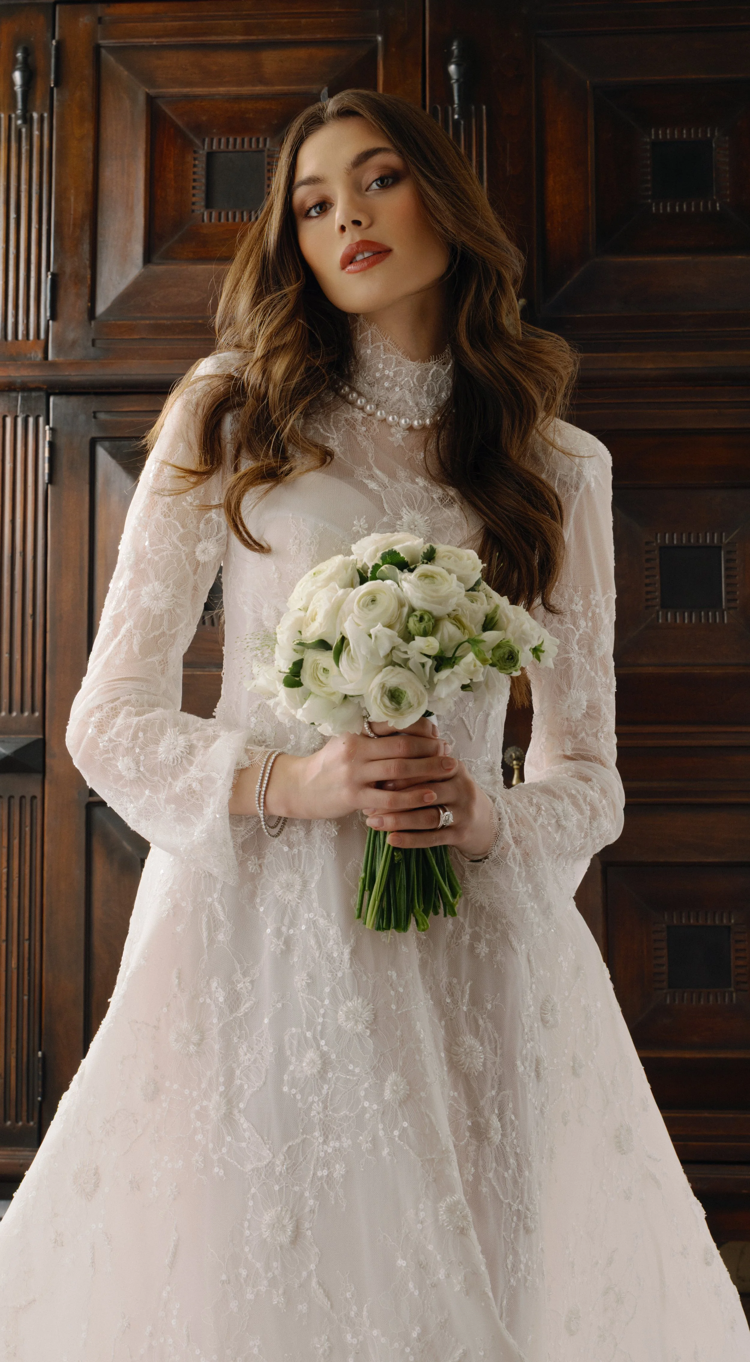A bride in a lace wedding dress holding a bouquet of white roses and greenery, standing in front of wooden paneling.