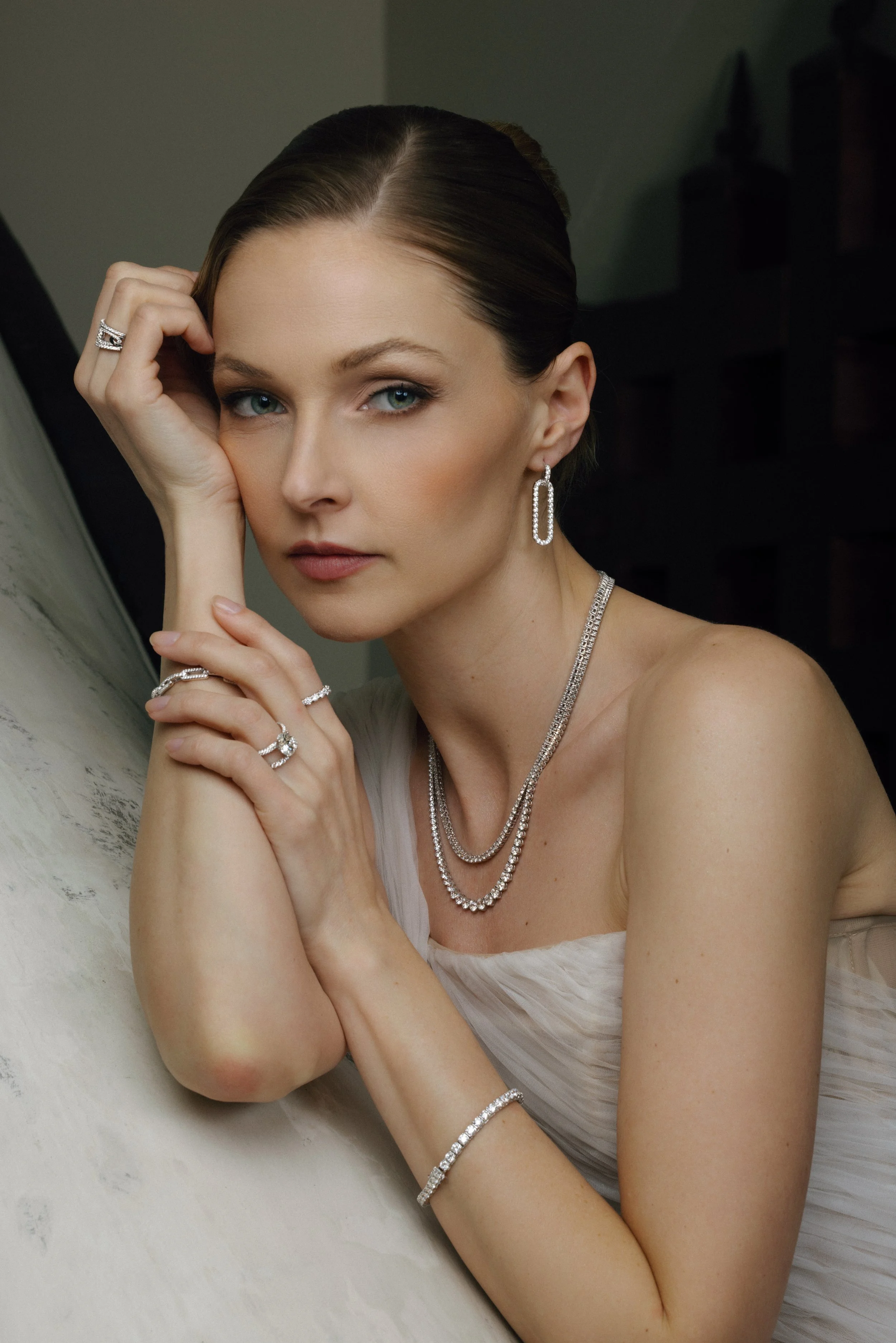 A woman wearing elegant diamond jewelry, including earrings, necklaces, rings, and a bracelet, posing with her head resting on her hand.
