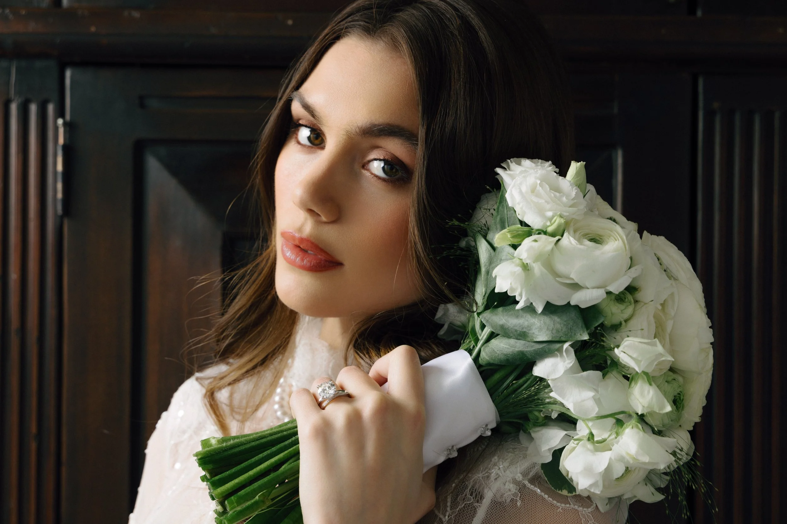 A woman with brown hair and jewelry holding a bouquet of white flowers, looking at the camera.