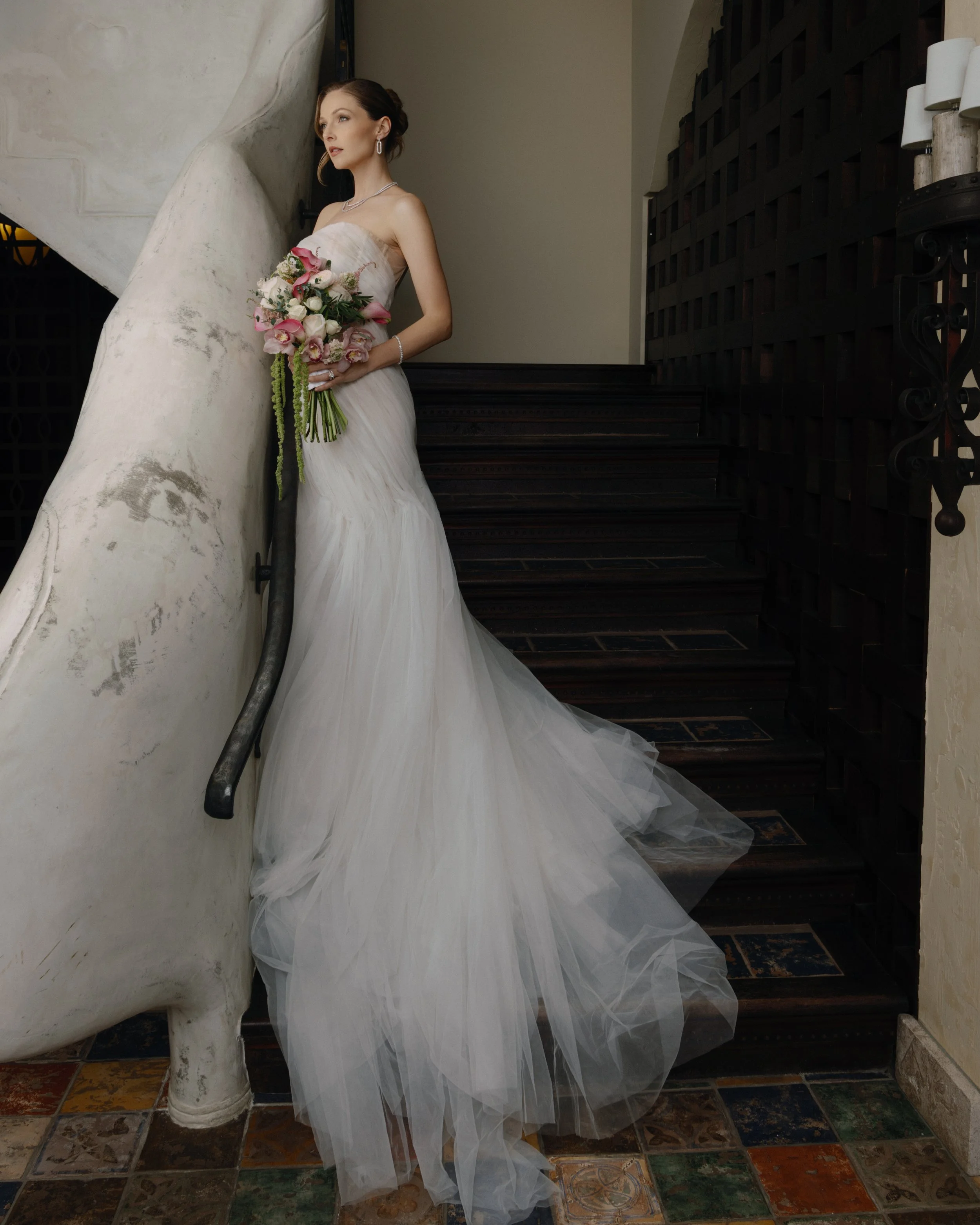 A bride in a white wedding dress holding a bouquet of pink and white flowers, standing on a staircase with dark wooden steps and a curved, textured wall.