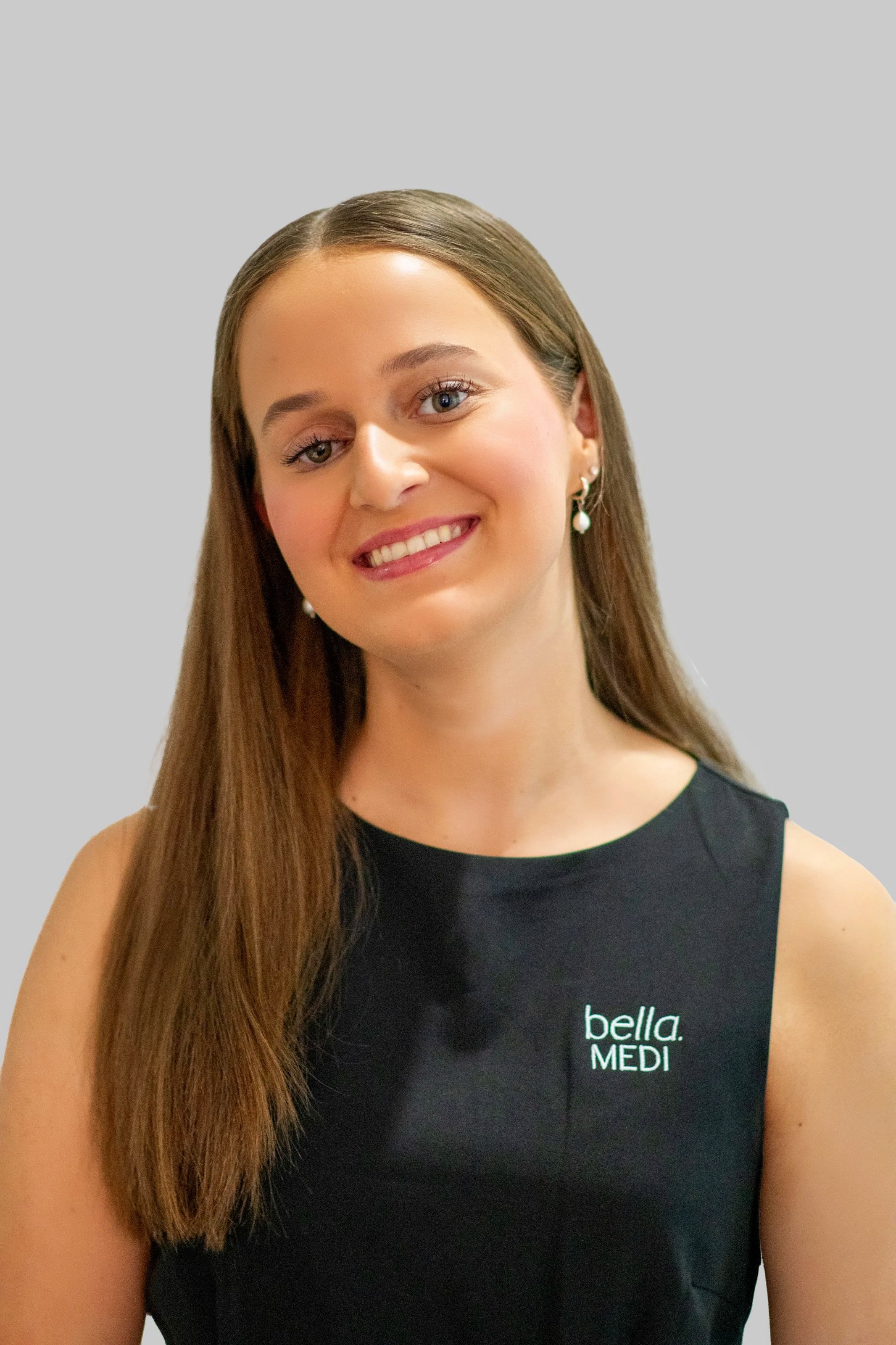 A skincare professional performing a facial treatment on a client, with the client lying down and wearing a white headband. The professional is wearing gloves and using a handheld device on the client's face in a clinic setting.
