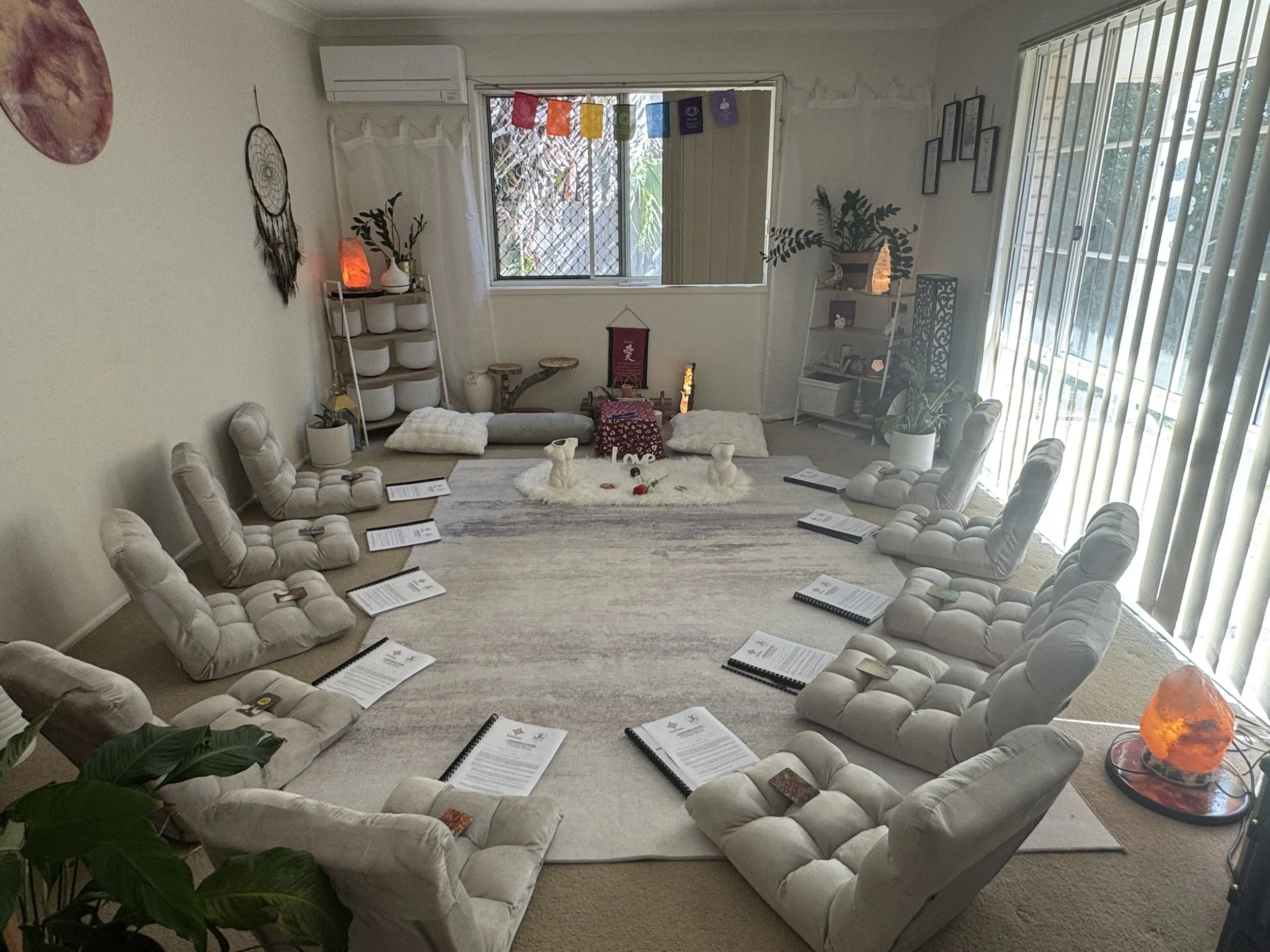 A room set up for tantra training, meditation or spiritual gathering with a circle of white cushioned chairs, each with a paper or booklet on the seat. In the center of the circle is an altar with rose petals and small candles.