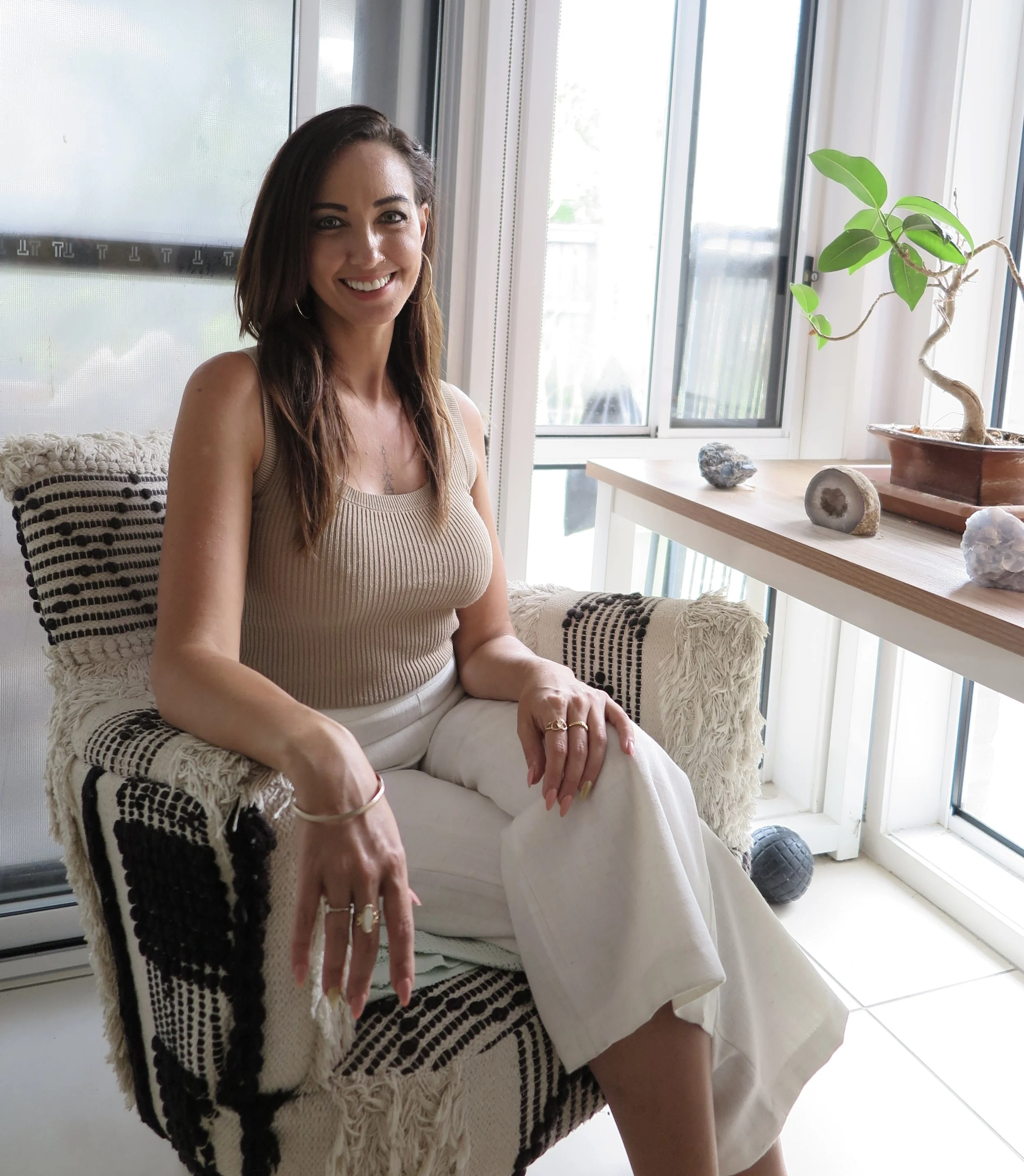A woman with long dark hair and hoop earrings sitting on a patterned armchair near large windows, with a wooden shelf holding a potted plant and rocks.