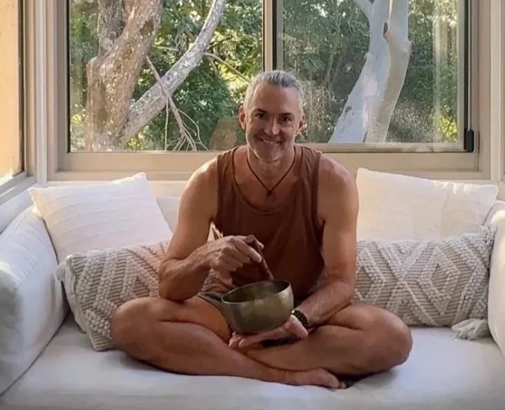 A man, Cass Jones, sitting cross-legged on a white couch, holding a singing bowl, smiling, with a large window behind him showing trees outside.