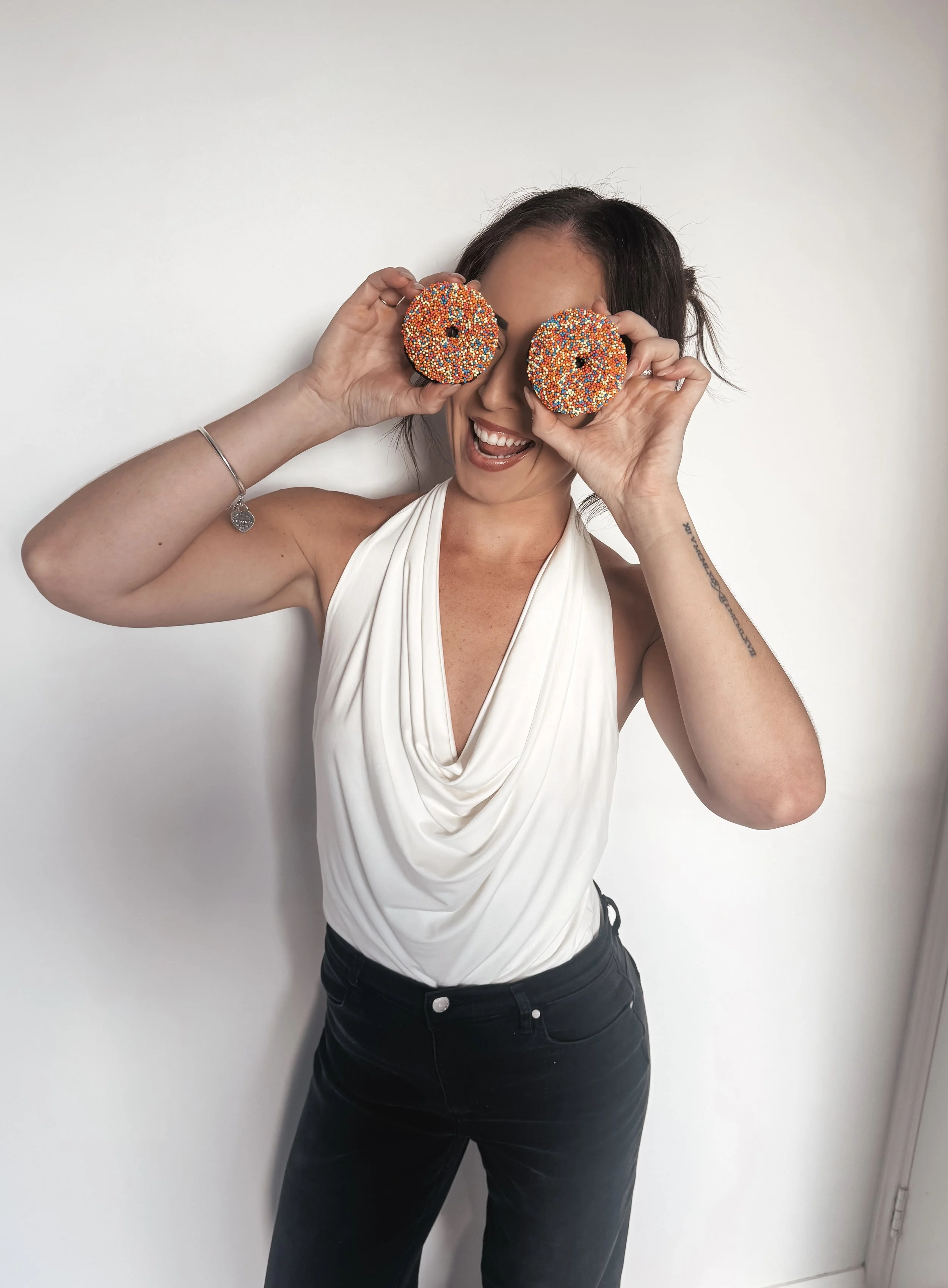 Woman smiling and holding two colorful donuts with sprinkles in front of her face, standing against a plain white wall.