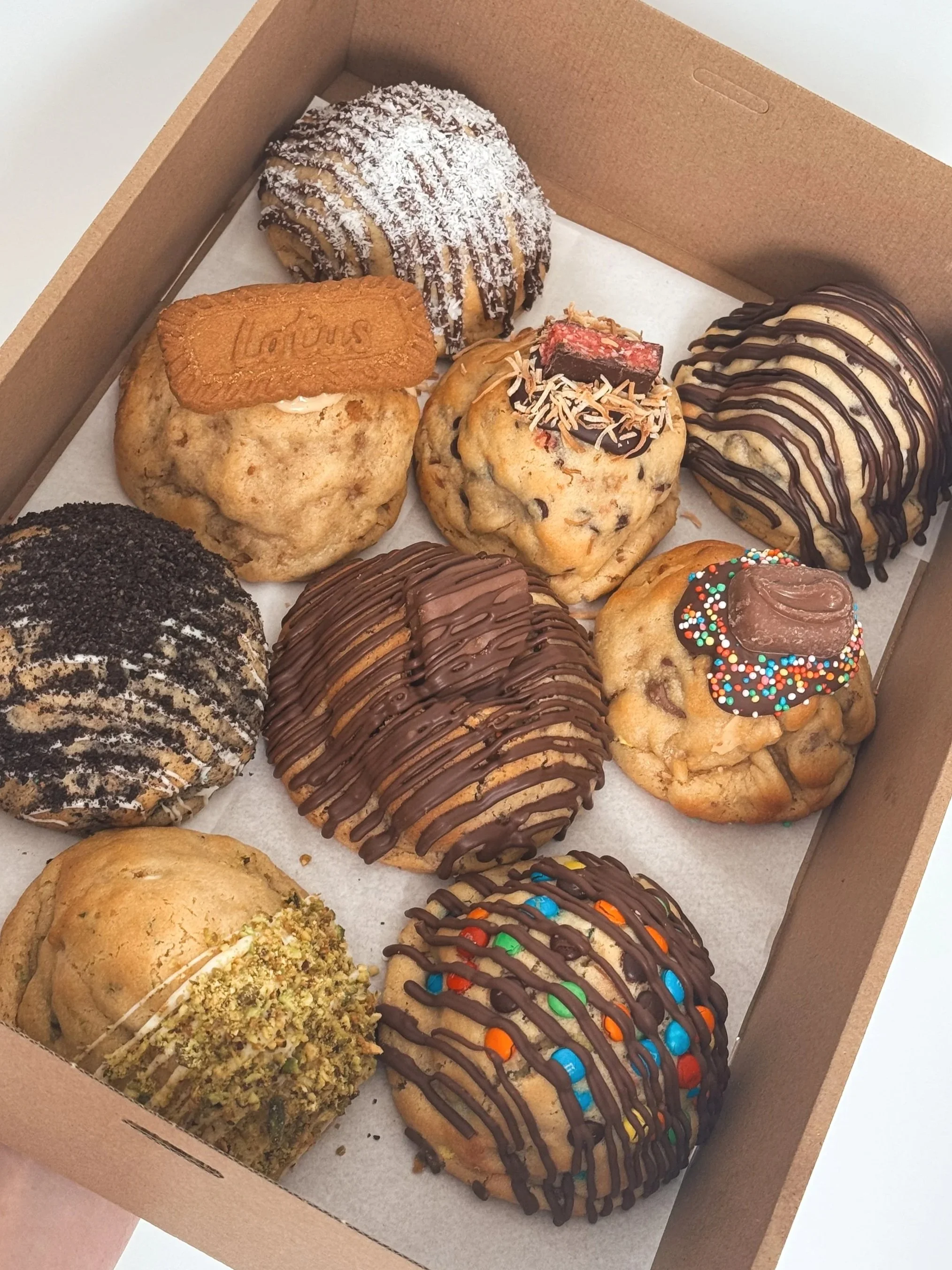 A cardboard box filled with an assortment of decorated cookies, including chocolate drizzled cookies, cookies with sprinkles, and cookies topped with crushed nuts.