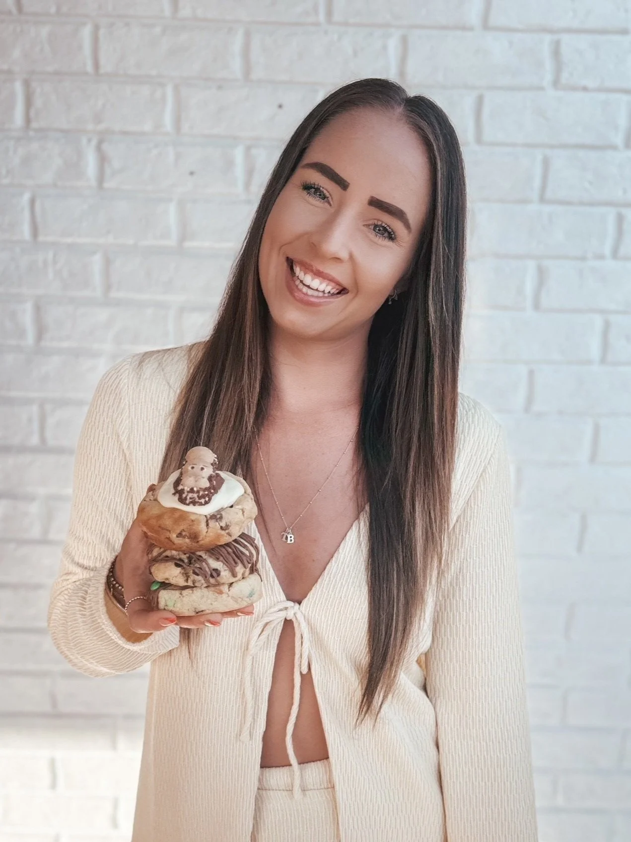A smiling woman with long dark hair and eyebrows holding a stack of four large cookies topped with a doll. Background is a white brick wall.