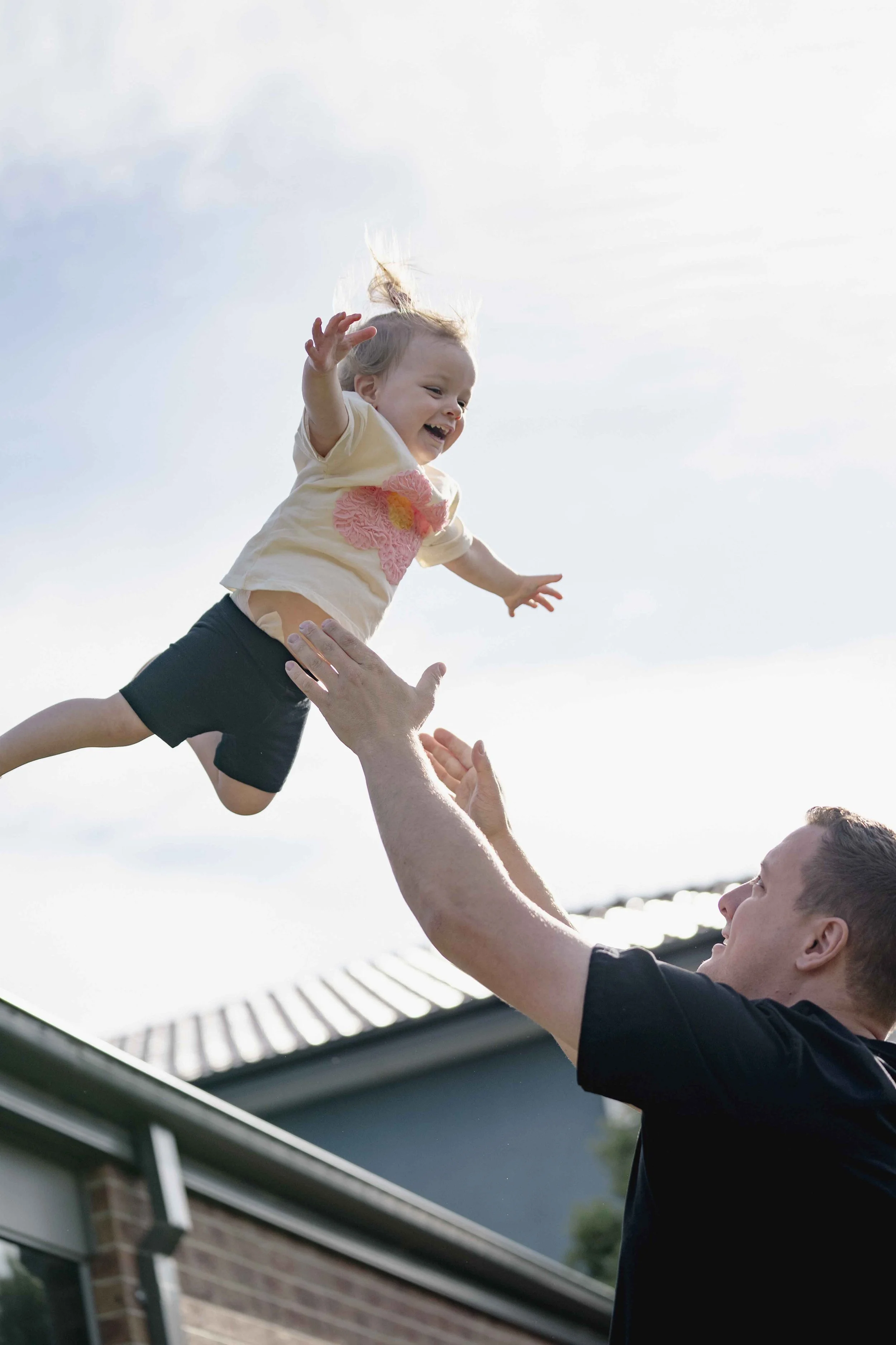 Dad tosses his toddler daughter into the air, capturing her laughter and pure joy. This candid family photography moment by Lowe & Co Photography highlights authentic connection, genuine smiles, and the beautiful chaos of real family life natural, he