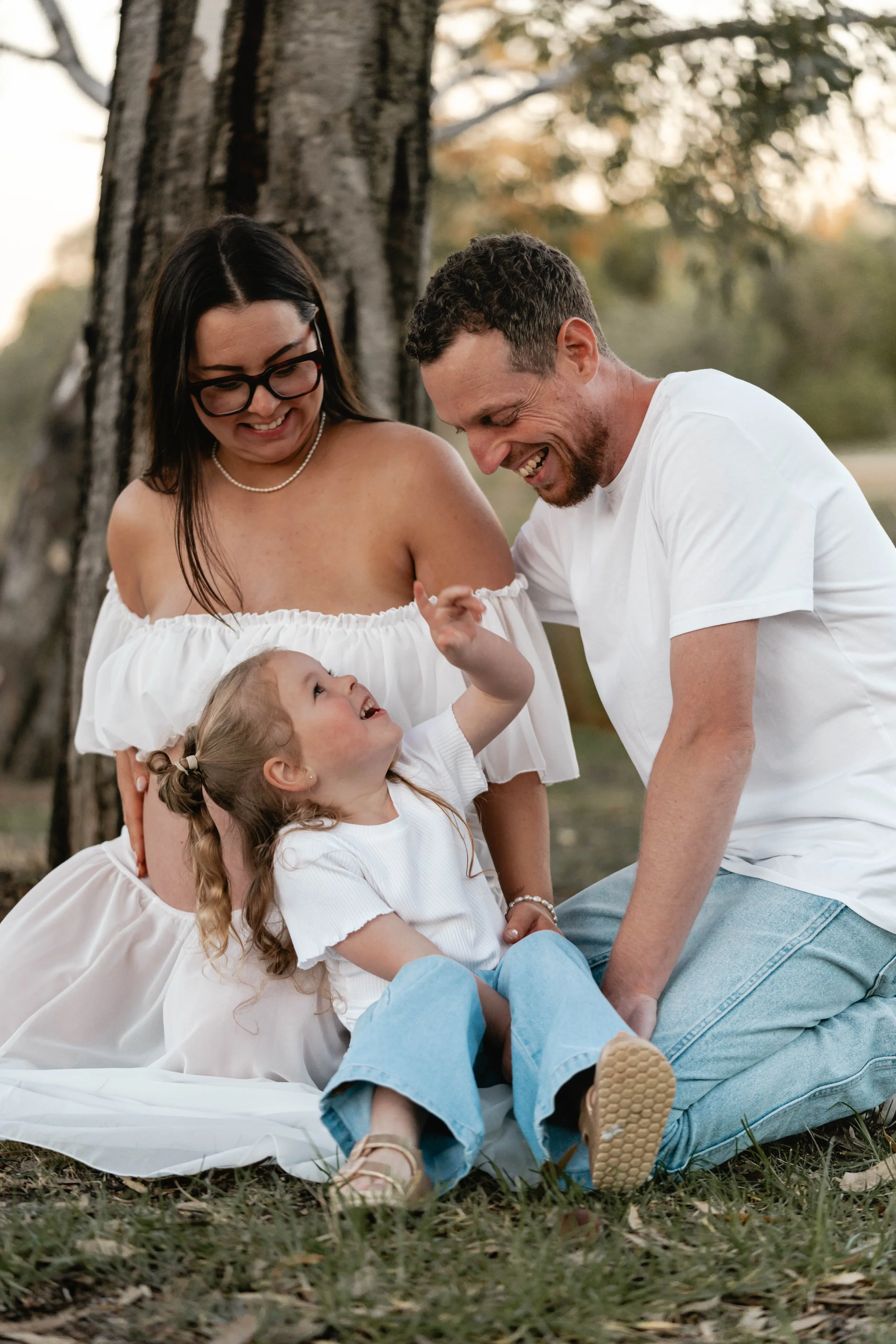 Family sitting under a tree, laughing together while their young daughter looks up with joy. This candid family photography moment by Lowe & Co Photography captures authentic connection, genuine smiles, and the heartfelt, timeless beauty of real fami