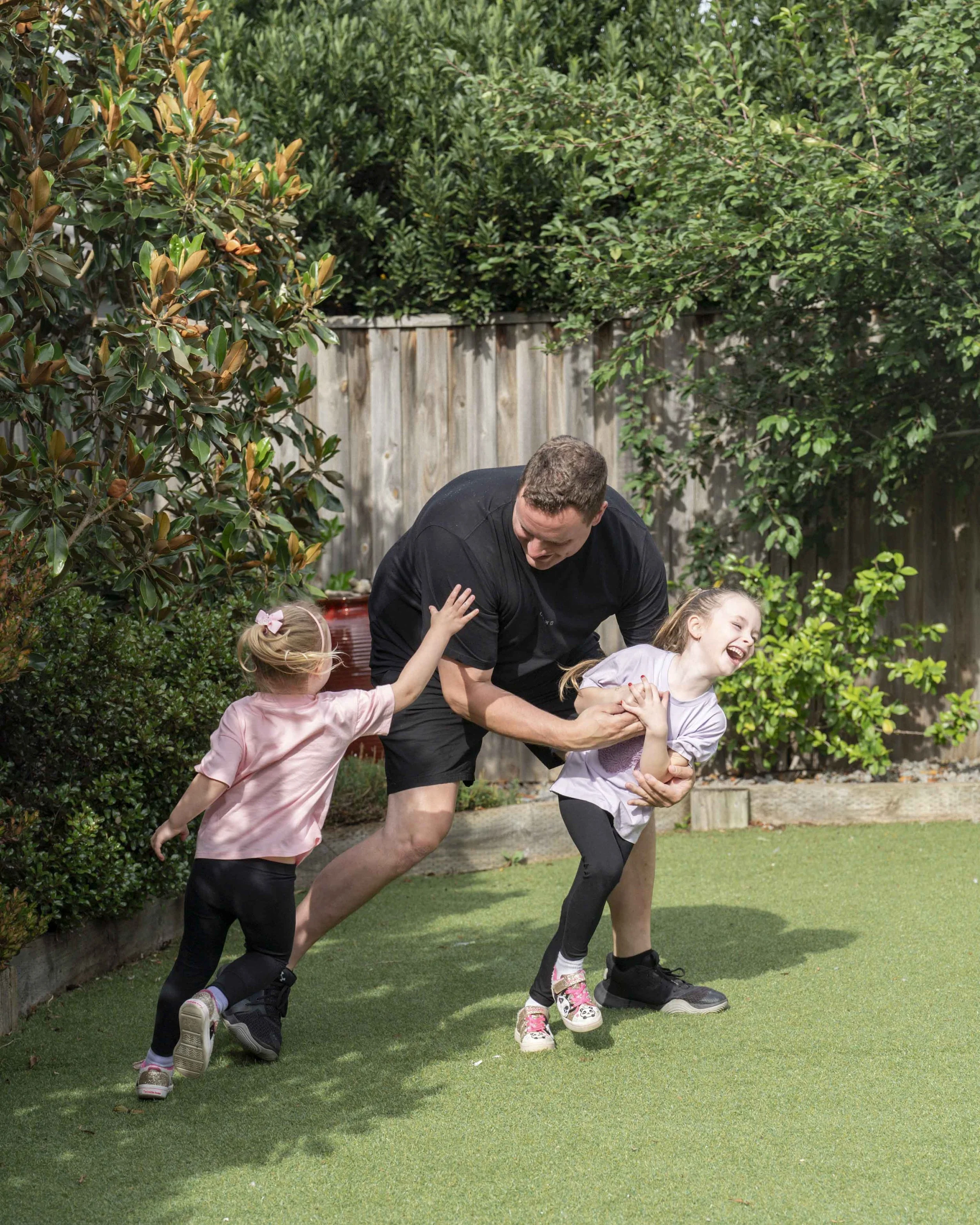 Father playing in his backyard with his two daughters. Running around with them tickling them. Genuine smiles and connection between them all. 