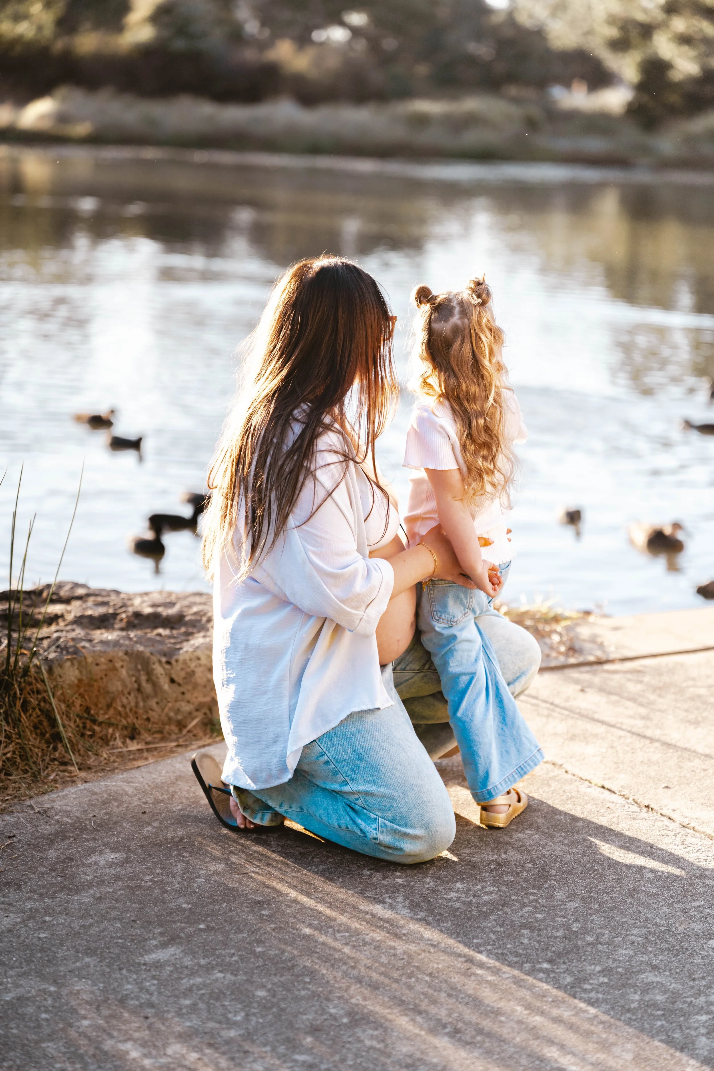 Mum kneels by the lake with her young daughter, watching ducks swim in the golden afternoon light. This candid family photography moment by Lowe & Co Photography captures authentic connection, natural beauty, and the timeless bond between parent and 