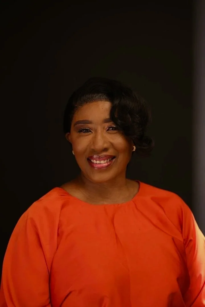 Portrait of Bukky Onifade, Founder Coached by Bukky smiling, wearing an orange top, with styled black hair, earrings, and makeup, against a dark background.