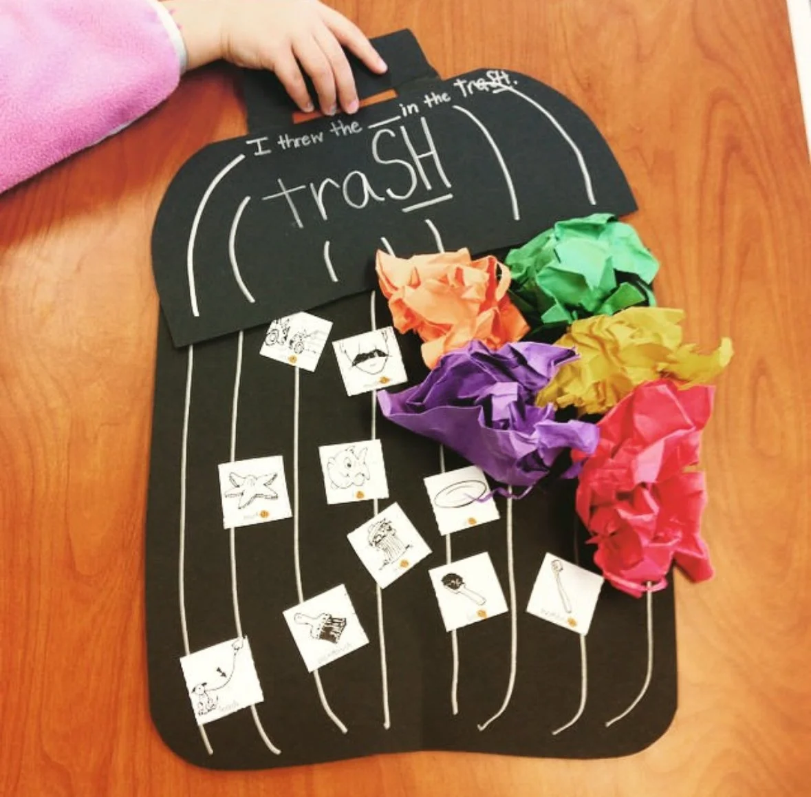 A classroom craft activity with a black paper tray labeled 'trash' and small paper bags of crumpled colored tissue paper in orange, green, purple, yellow, and red, representing trash items, with small pictures of trash items attached to white strings.