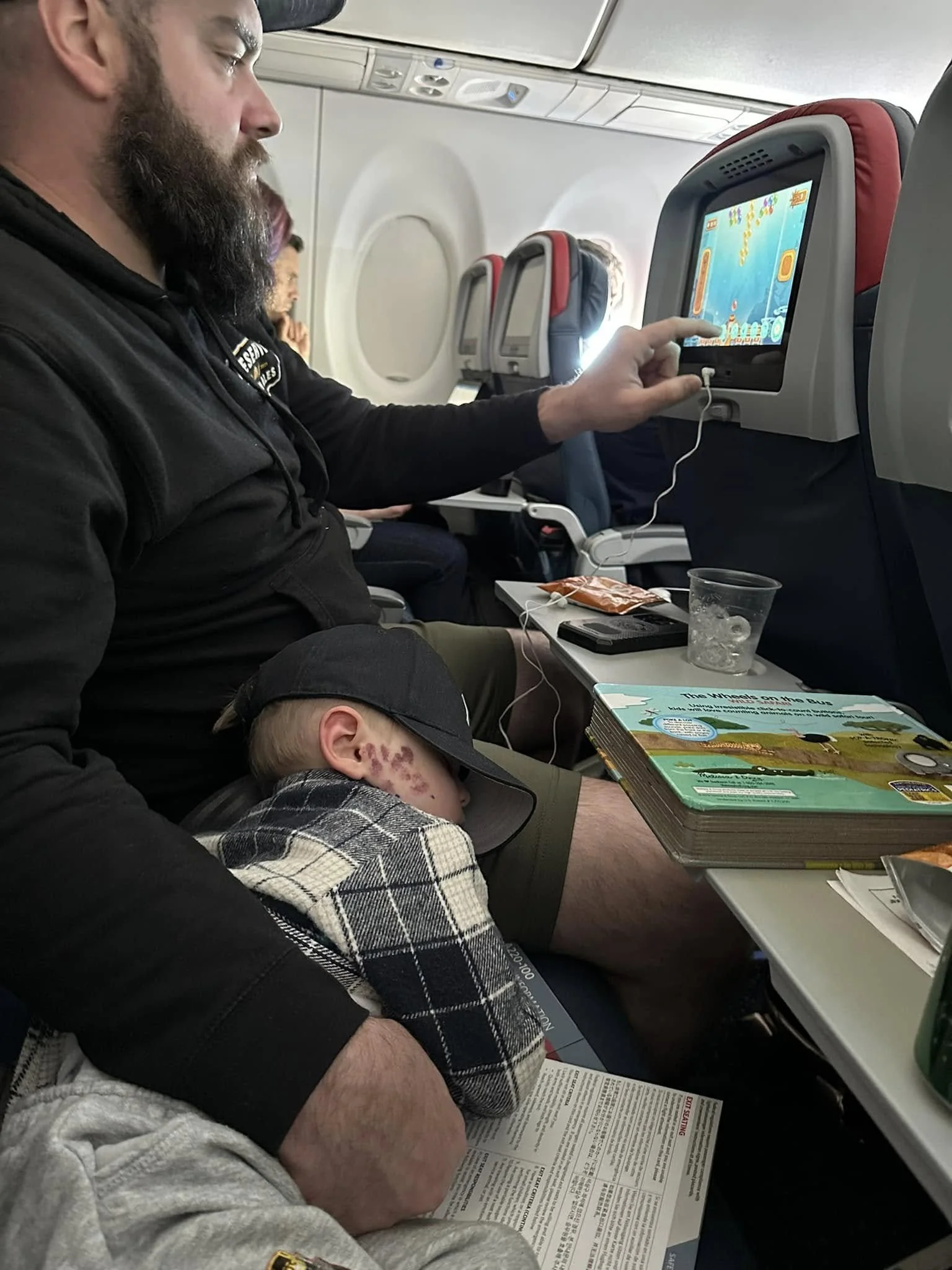 A man and a child sleeping on an airplane, with the man reading a newspaper and using a tablet. The child is wearing a hoodie with a checkered pattern and a black cap.