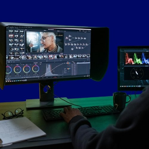 A person working on video editing on a computer with color grading tools, with a secondary monitor displaying audio frequency graphs, on a desk with a notebook, glasses, and a coffee mug.