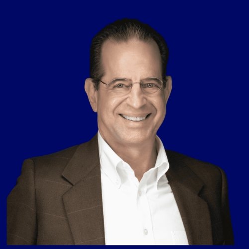 A man with glasses, dark hair, and a smile, wearing a dark blazer over a white shirt, against a dark blue background.