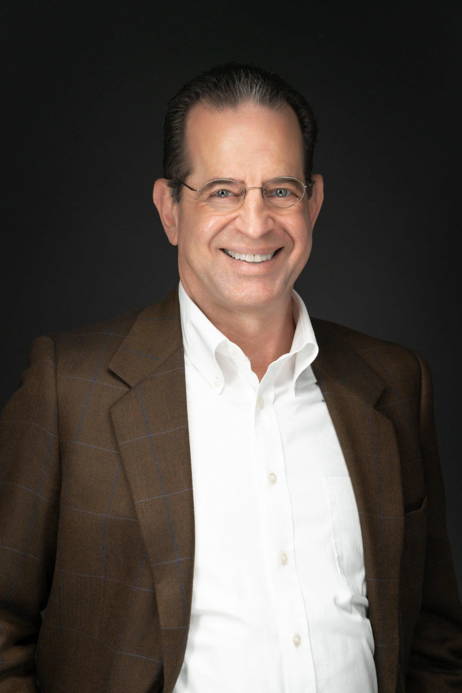 Headshot of a man with glasses smiling, wearing a brown blazer and white dress shirt against a black background.