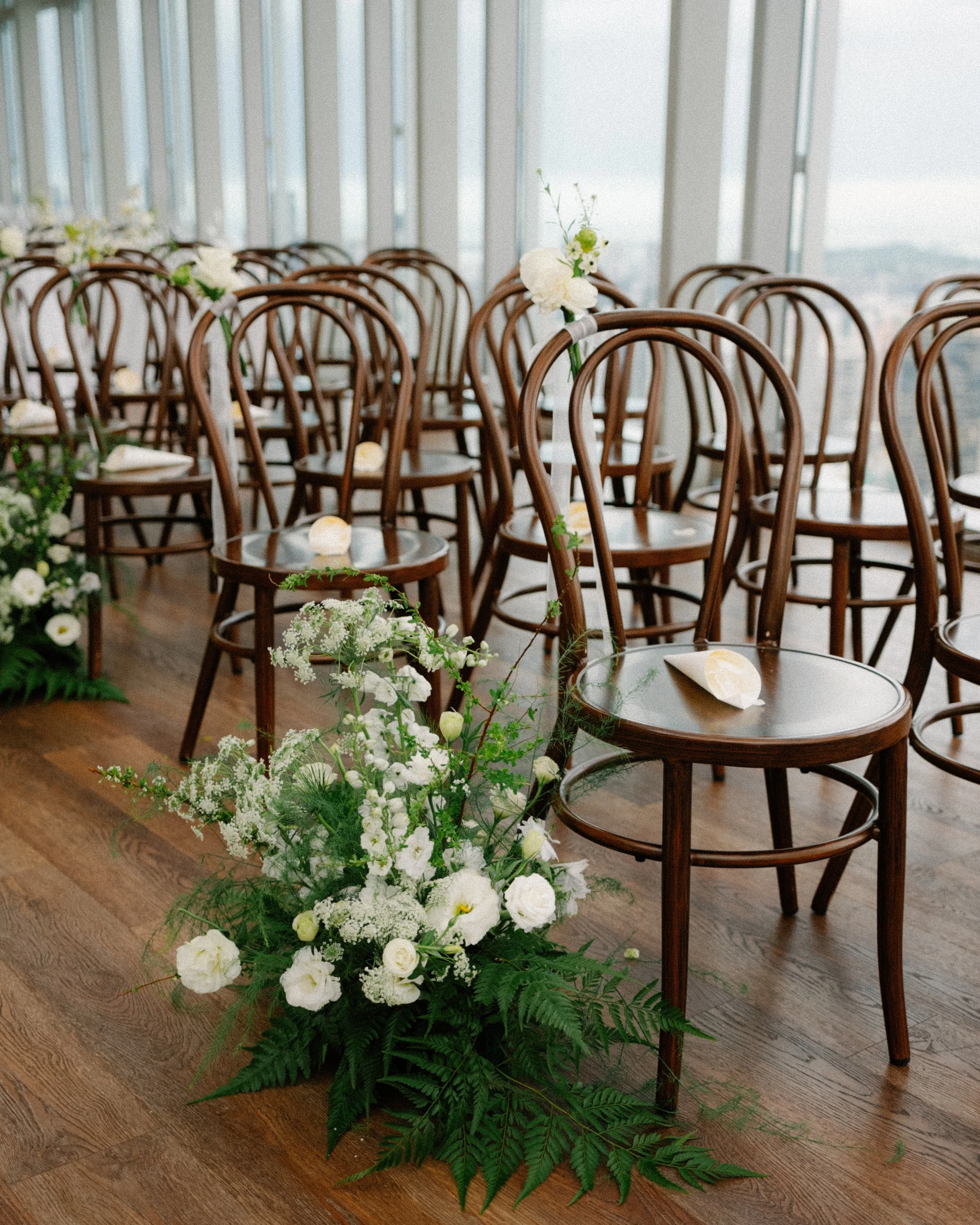 Wooden bentwood ceremony chairs with white wildflower aisle florals at 1-Atico Singapore wedding venue with floor-to-ceiling windows.