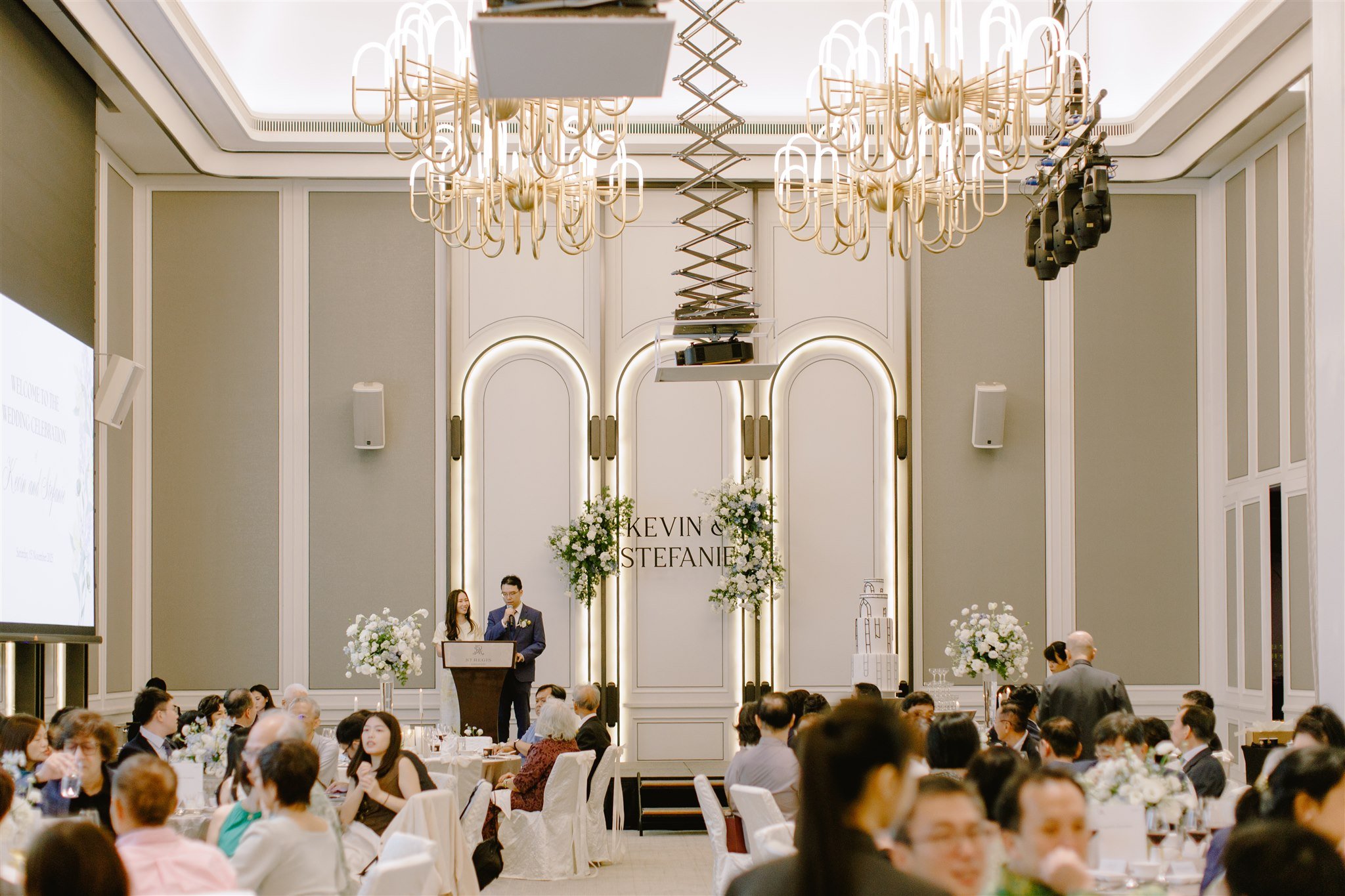 Wedding reception ballroom with elegant chandeliers, floral arrangements, and a personalised stage backdrop for Kevin and Stefanie’s celebration in Singapore.