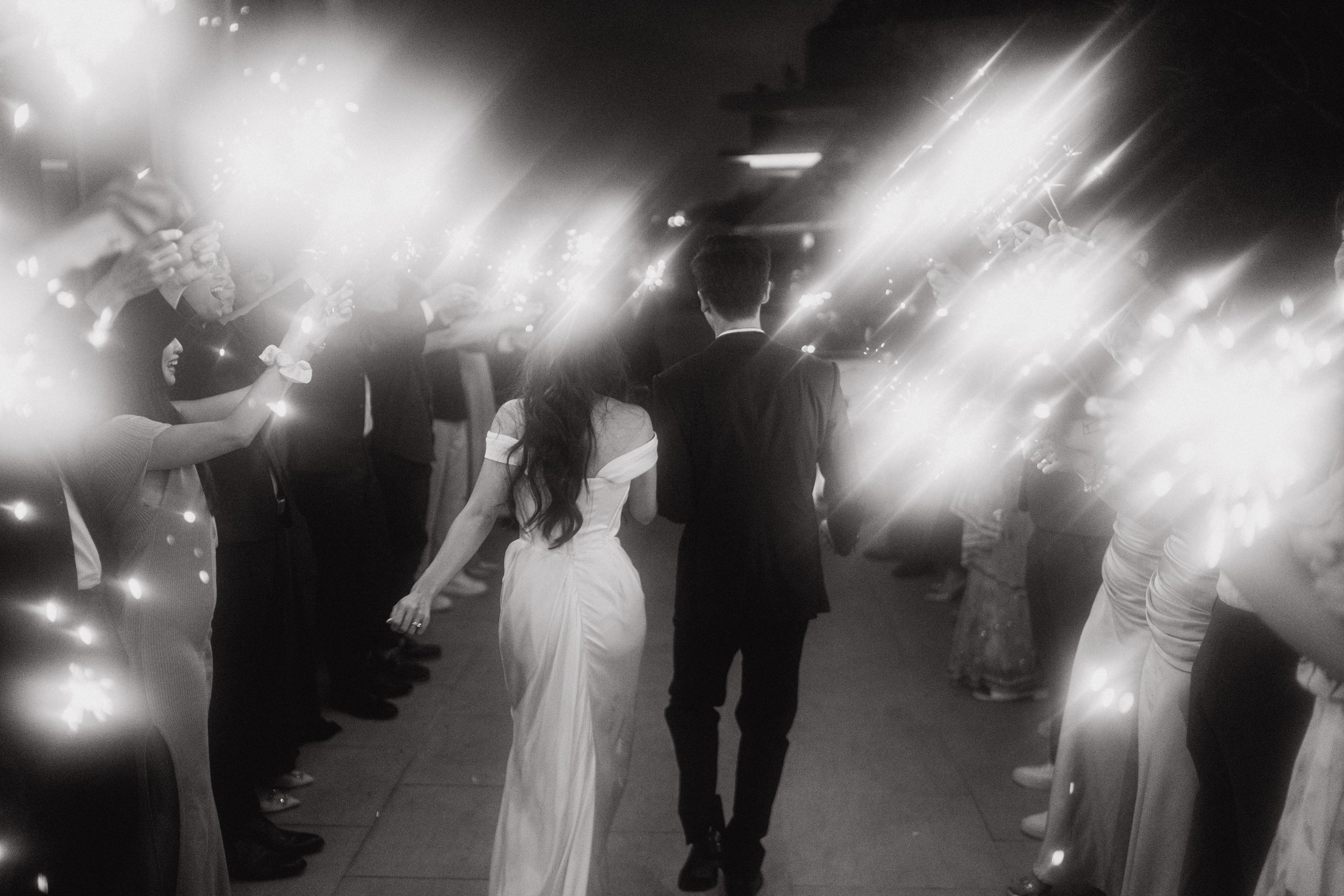 A black and white photo of a wedding reception, featuring a bride in a white off-shoulder gown and a groom in a dark suit, holding hands and walking through a corridor lined with guests who are holding sparklers.