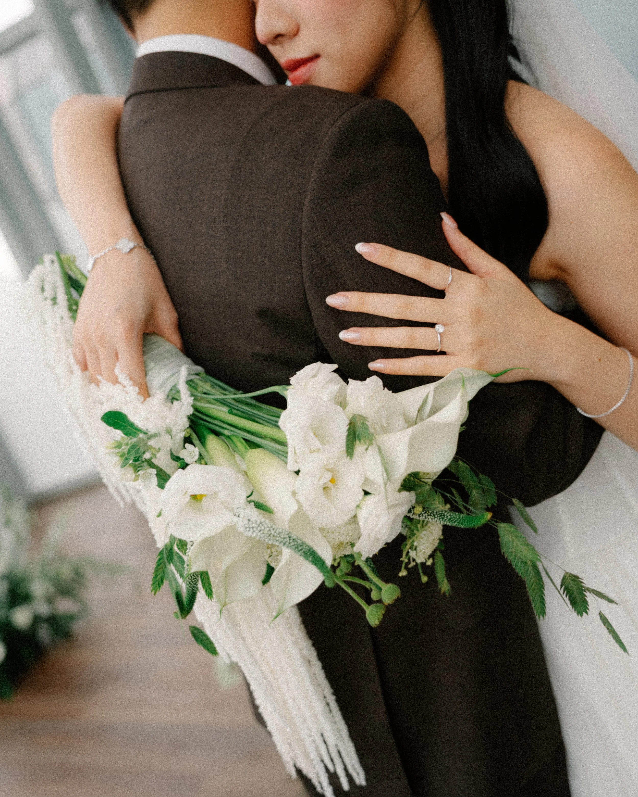 Close-up of bride embracing groom in brown suit with white calla lily and wildflower bouquet.